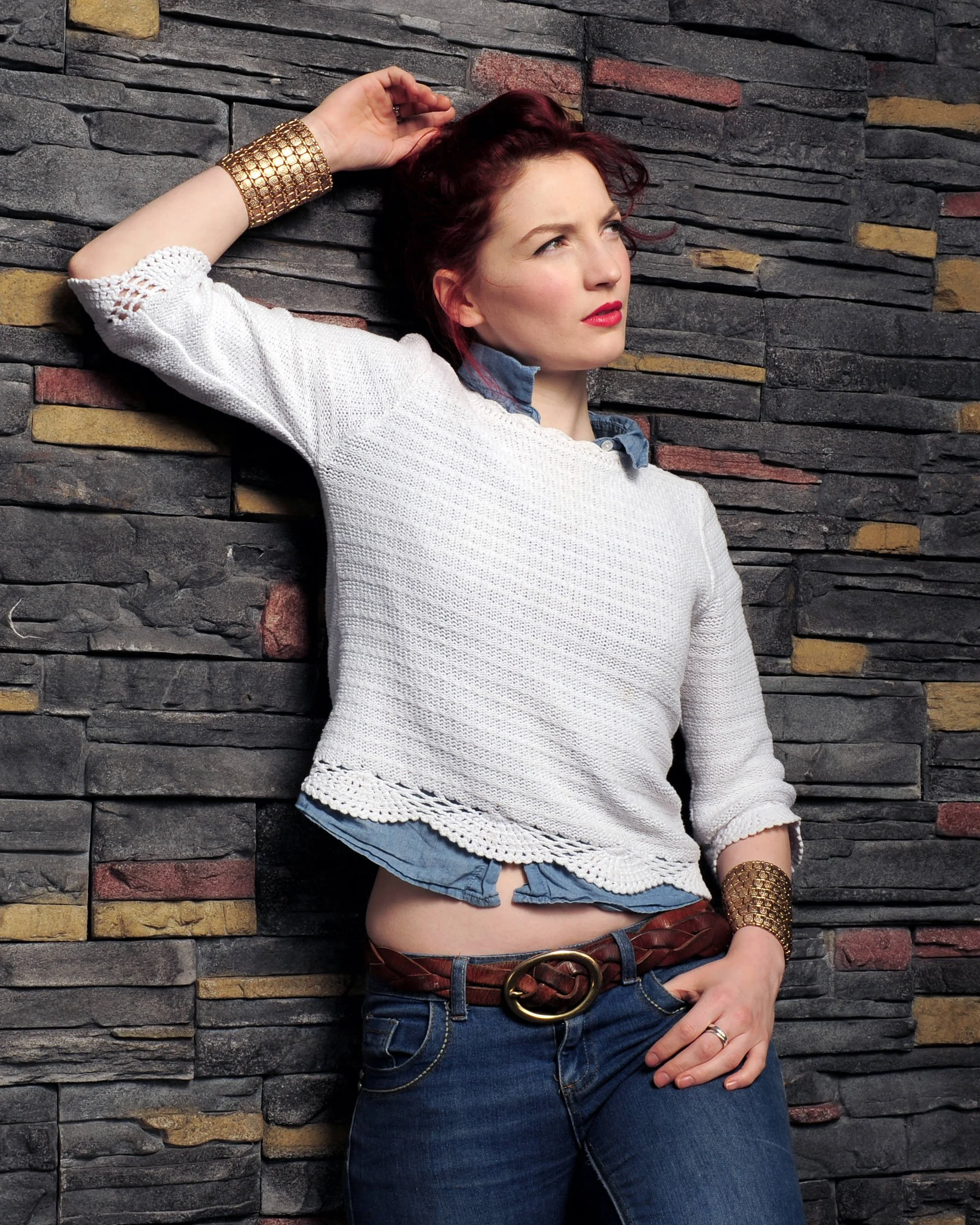 A woman with red hair and fair skin leaning against a stone wall, looking away with a thoughtful expression, wearing a white knit sweater over a blue shirt, jeans, a brown belt, and a wide gold cuff bracelet.