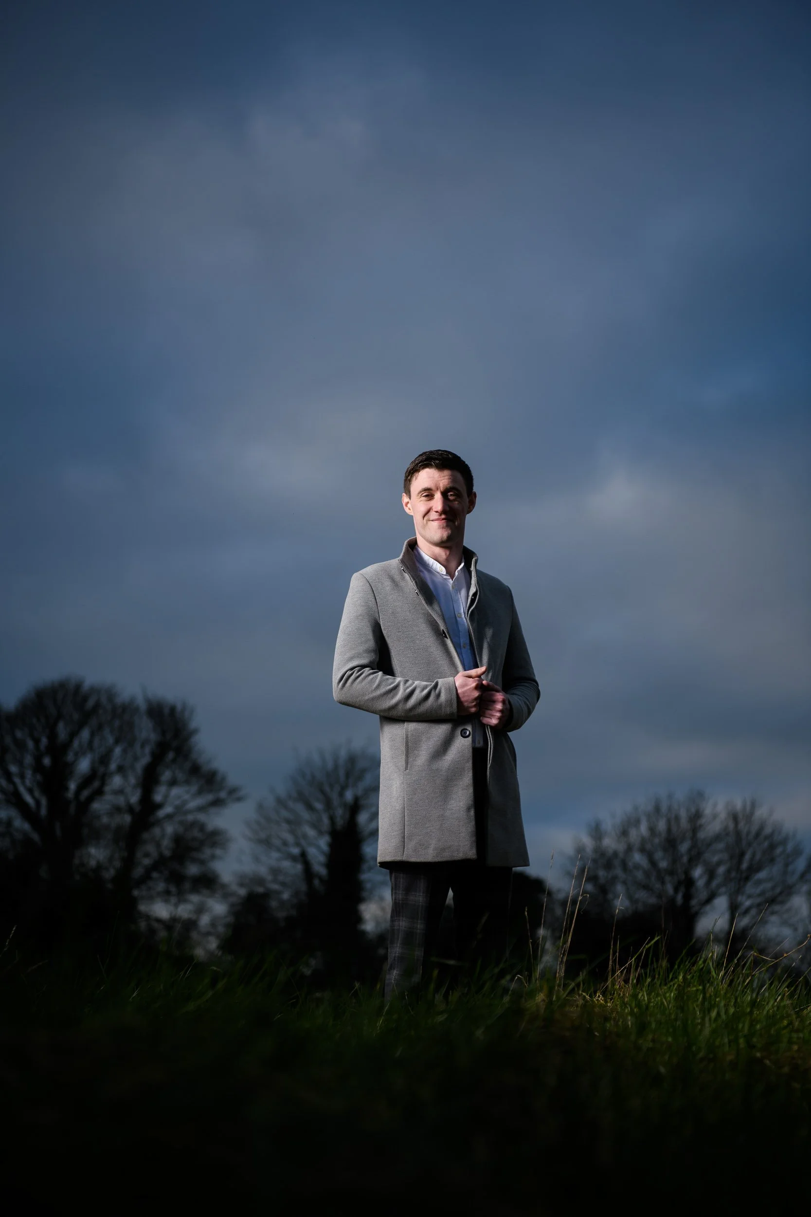 A man standing outdoors on a grassy field during overcast weather, wearing a gray coat and posing with a slight smile against a dark cloudy sky with trees in the background.