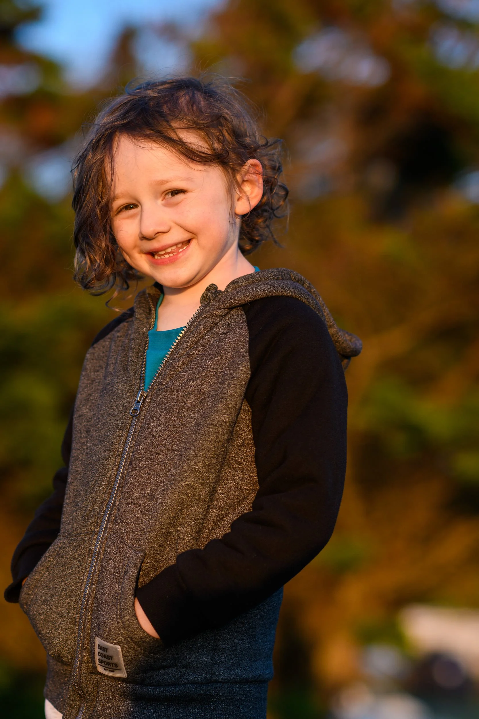 A young boy with curly brown hair smiling outdoors during sunset, wearing a gray and black zip-up jacket with his hands in his pockets.