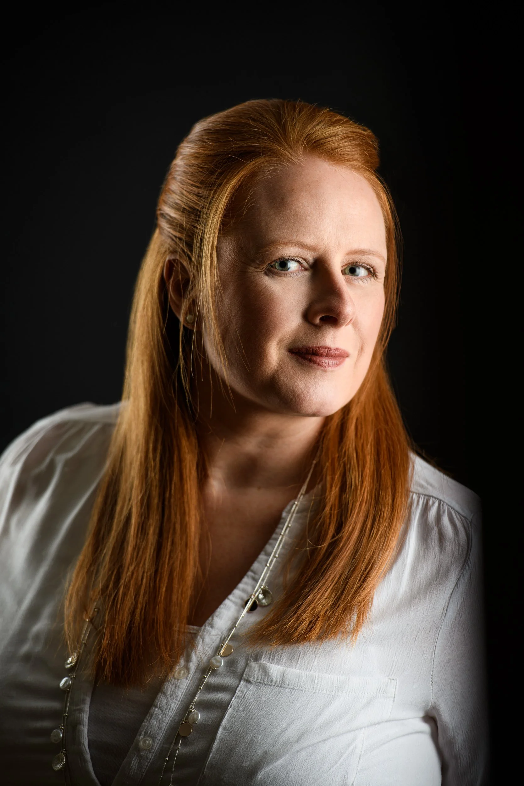 Portrait of a woman with long red hair styled back, wearing a white blouse and necklace, with a soft expression against a dark background.
