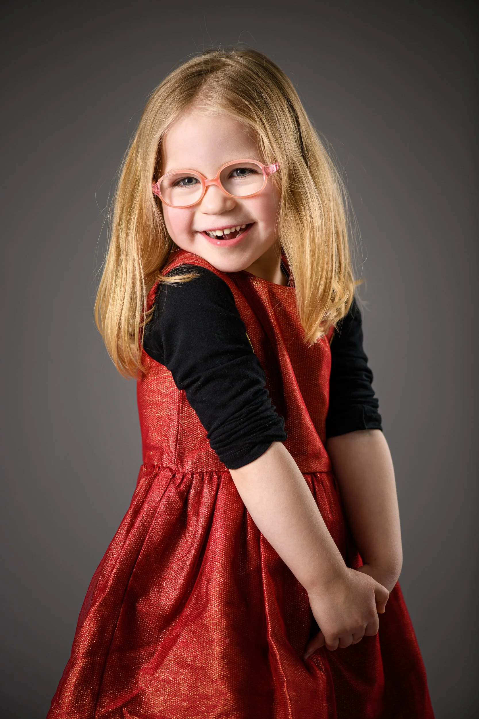 A young girl with red hair, wearing pink glasses, a red dress with a black long-sleeve shirt underneath, smiling and showing no teeth.