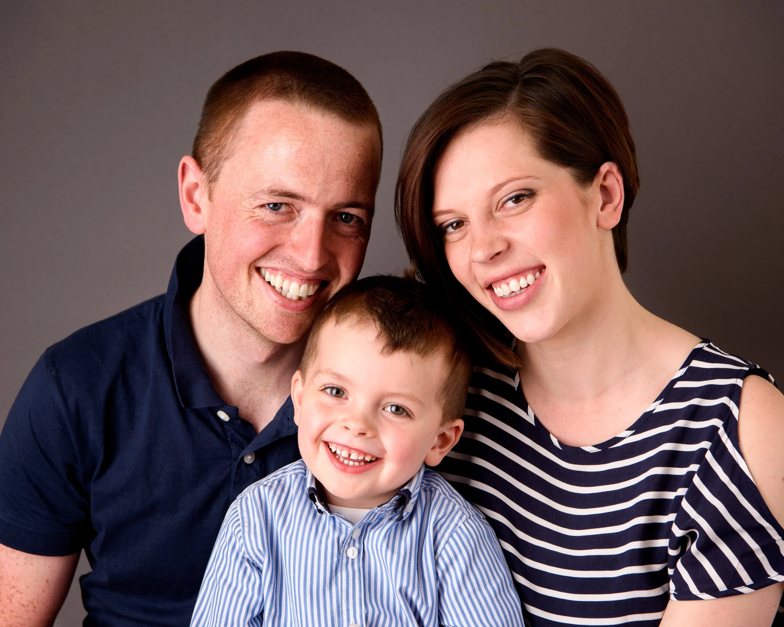 A smiling family of three posing together against a plain background.
