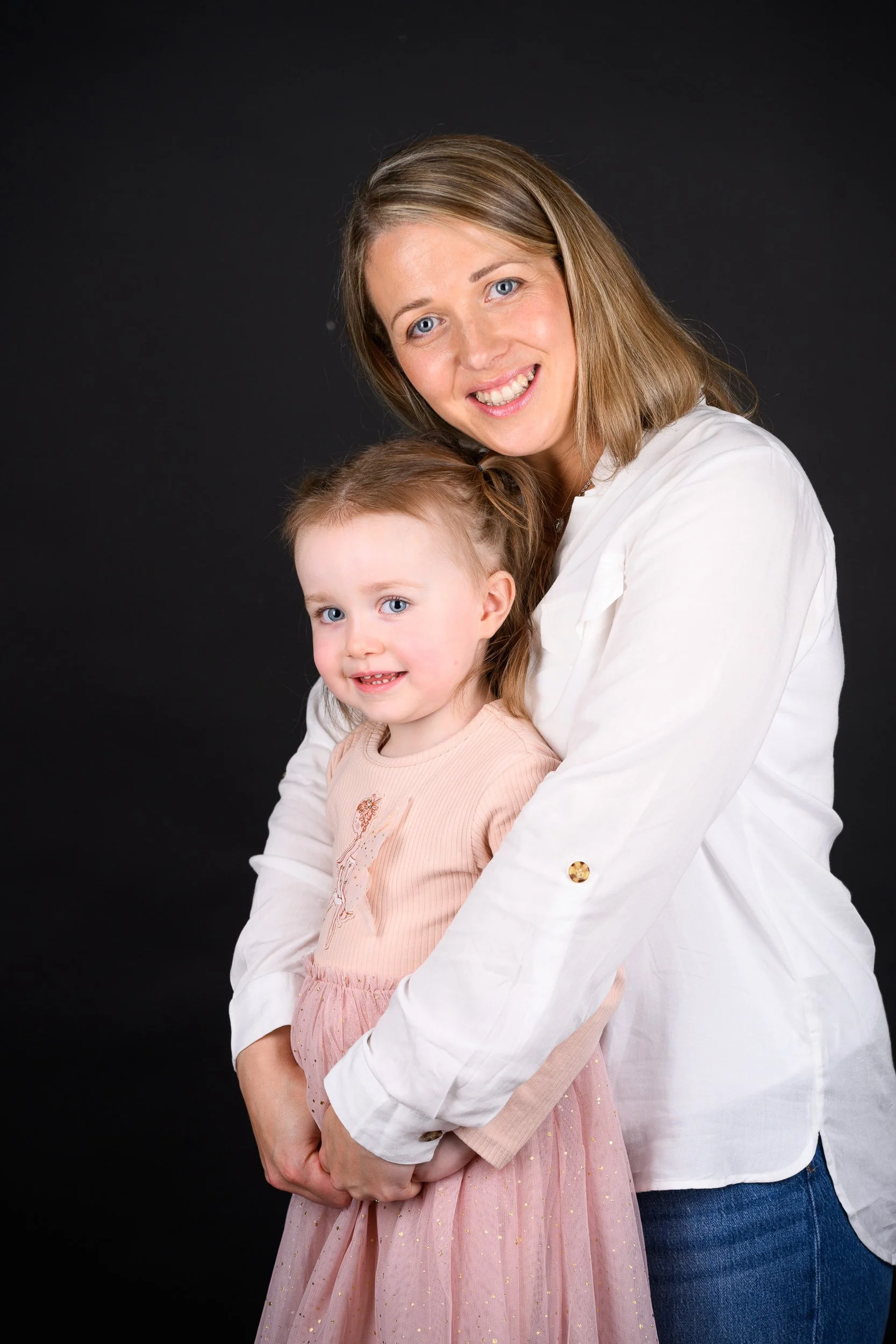 A woman with shoulder-length light brown hair and a young blonde girl smiling together against a dark background.
