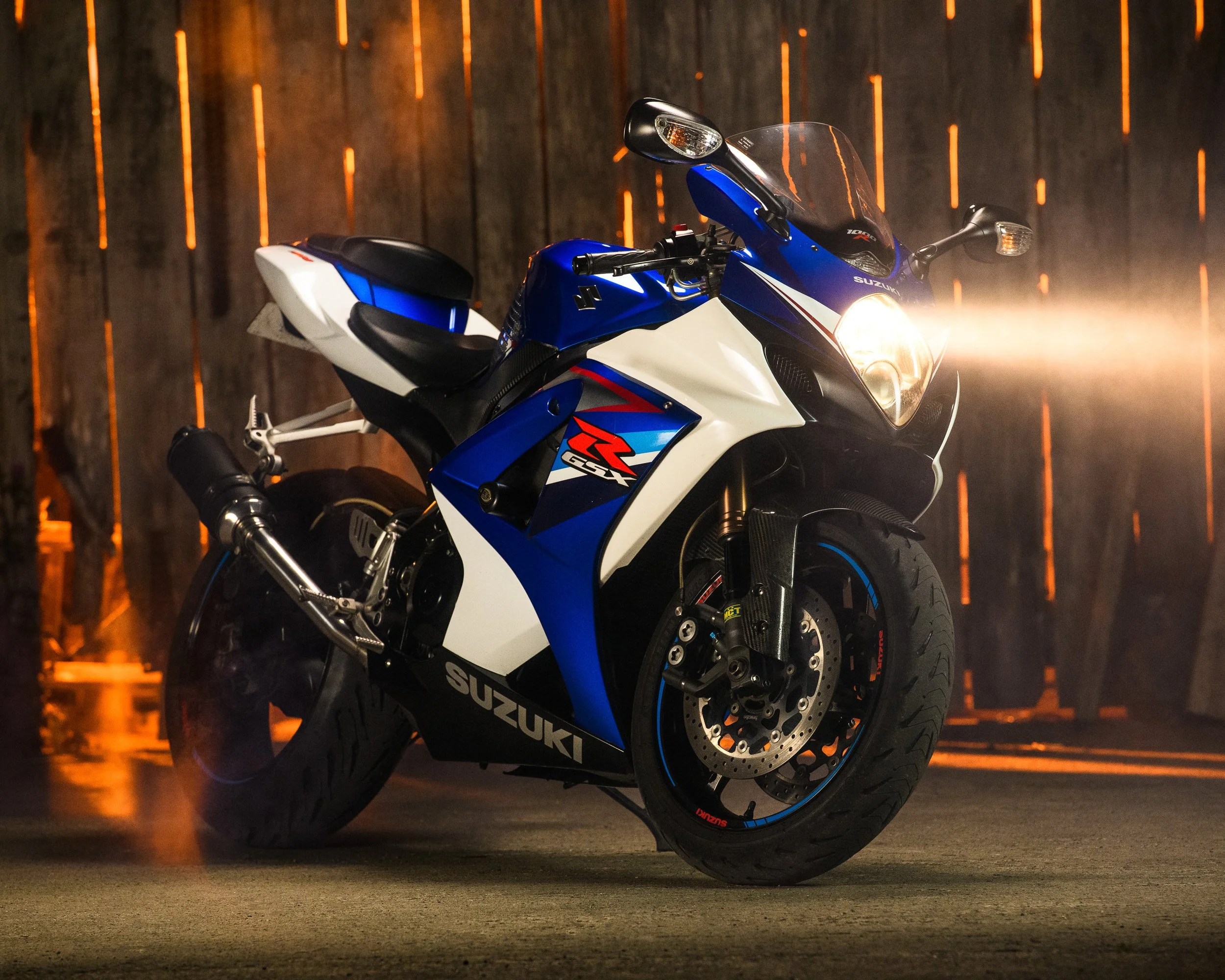 Suzuki GSX-R in blue, white and black, photographed indoors with dramatic lighting across the front and headlight illuminated.
