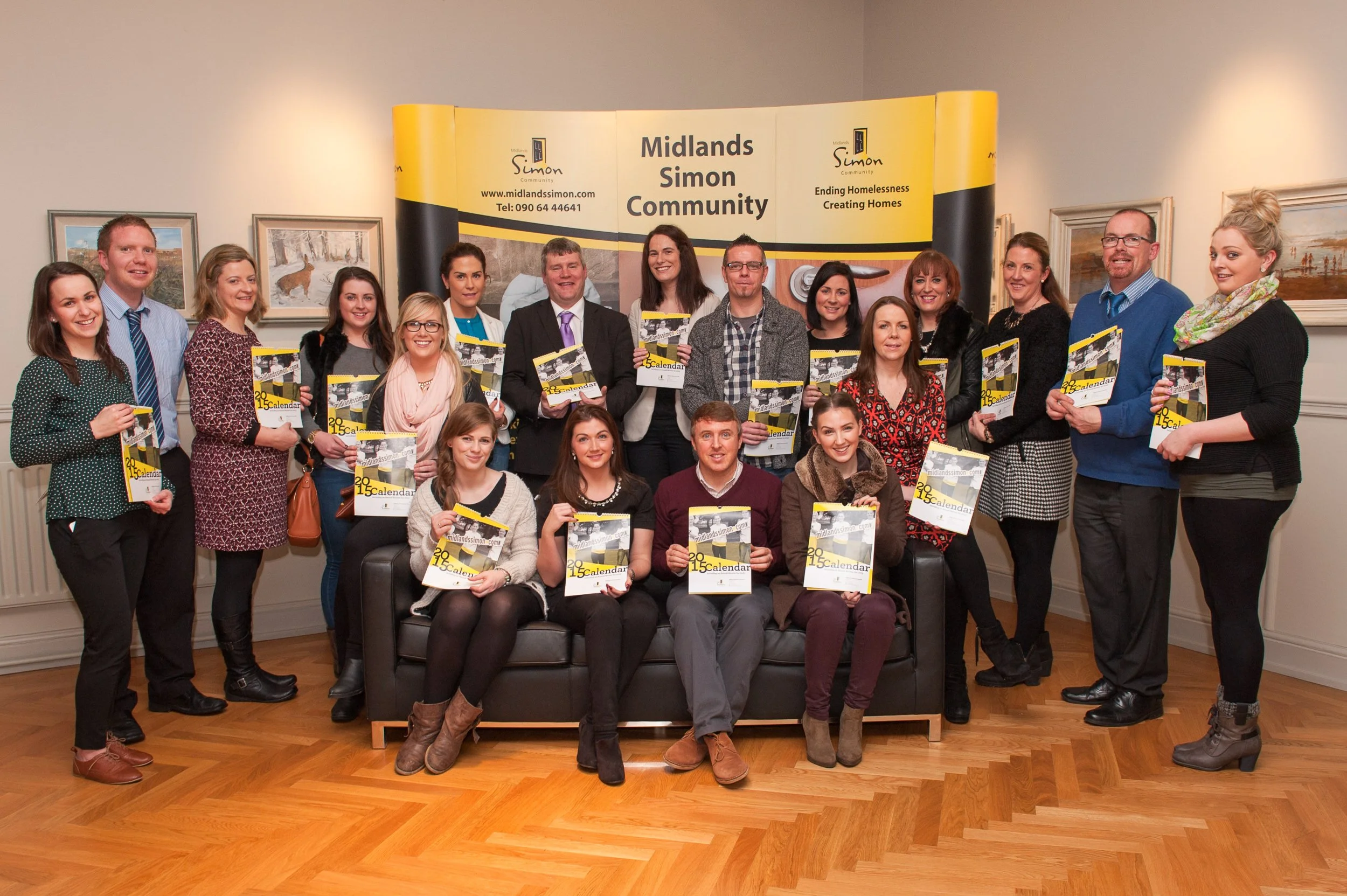 Group of people gathered at the Midlands Simon Community event, holding calendars, posing for a group photo in a room with framed pictures and a yellow and black banner in the background.