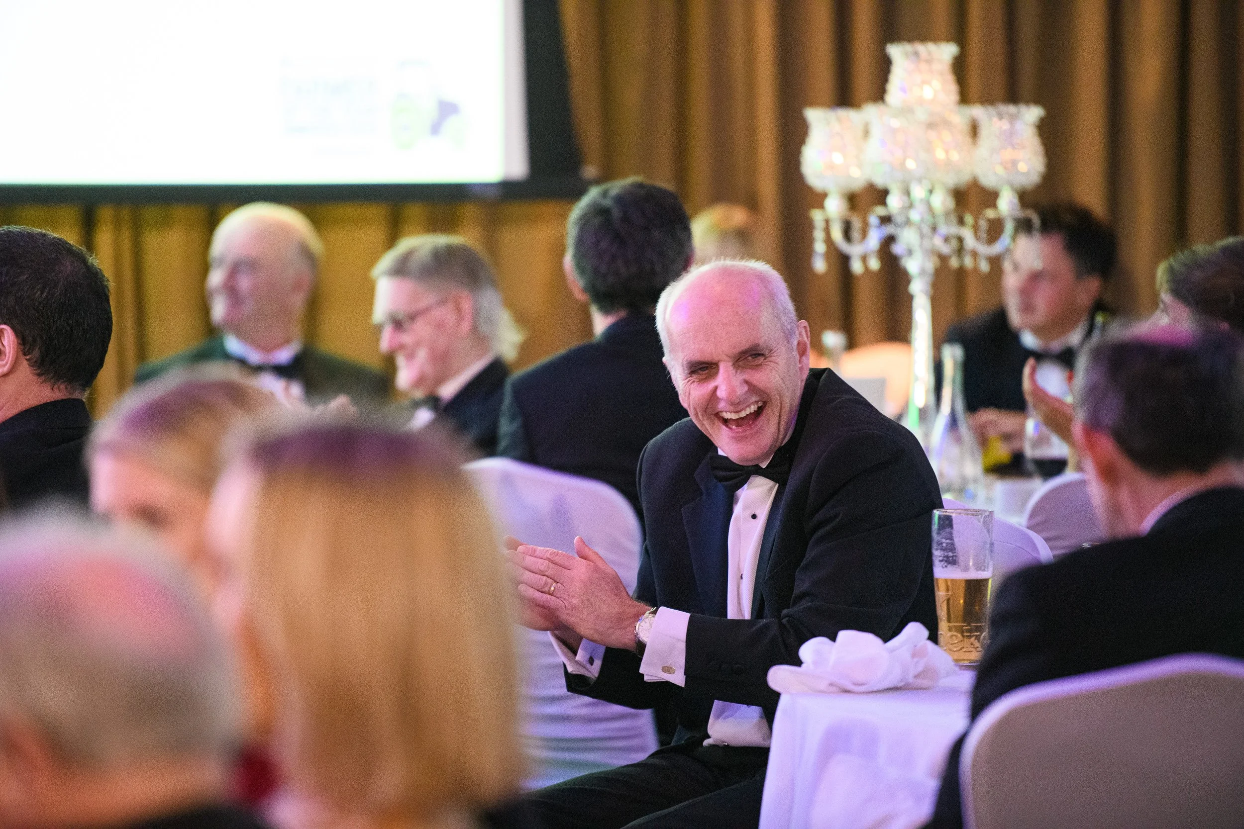 Men in tuxedos sitting at tables during a formal event, with one man in the foreground laughing and clapping.