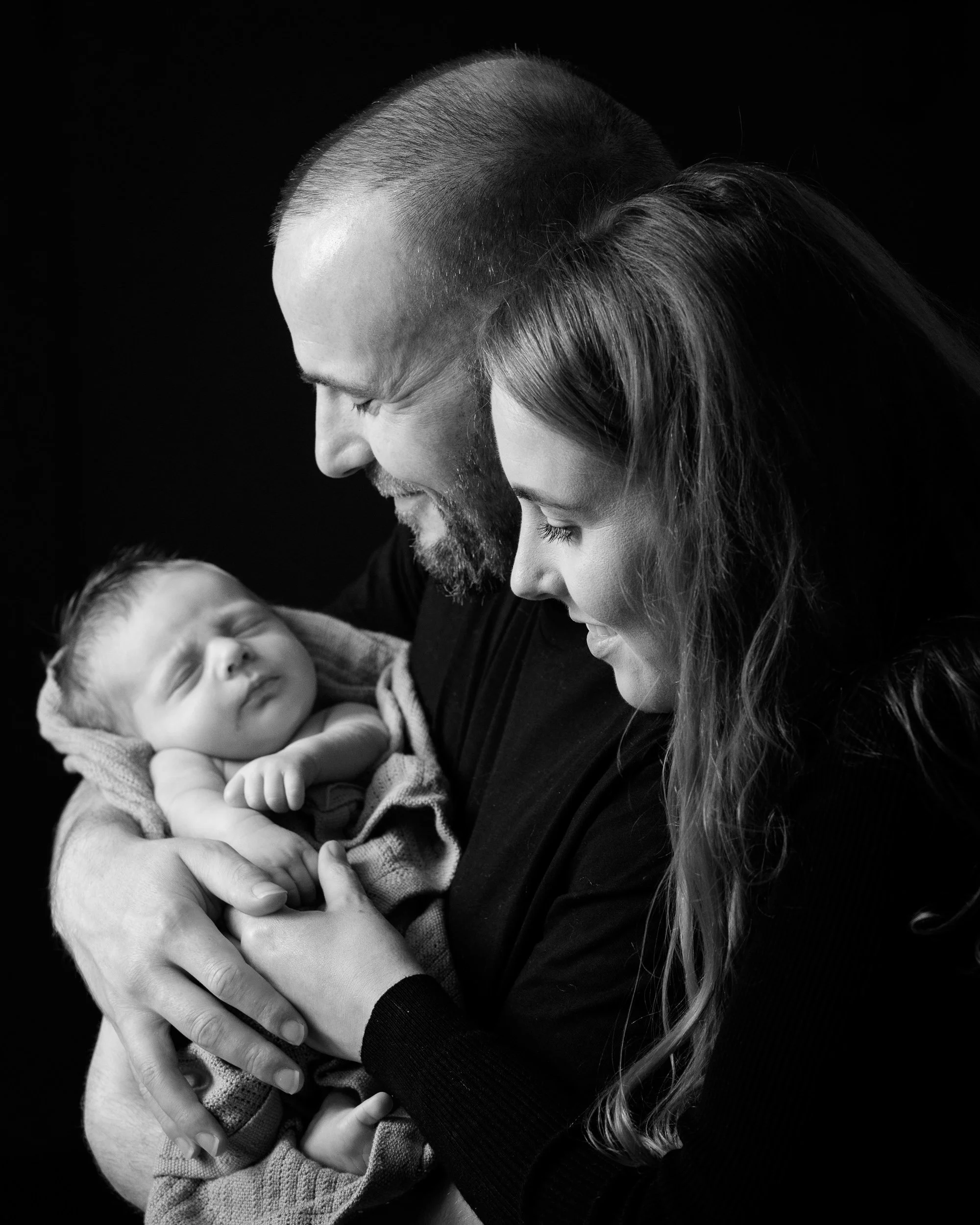 Black and white portrait of a couple holding their sleeping newborn, smiling at each other.

