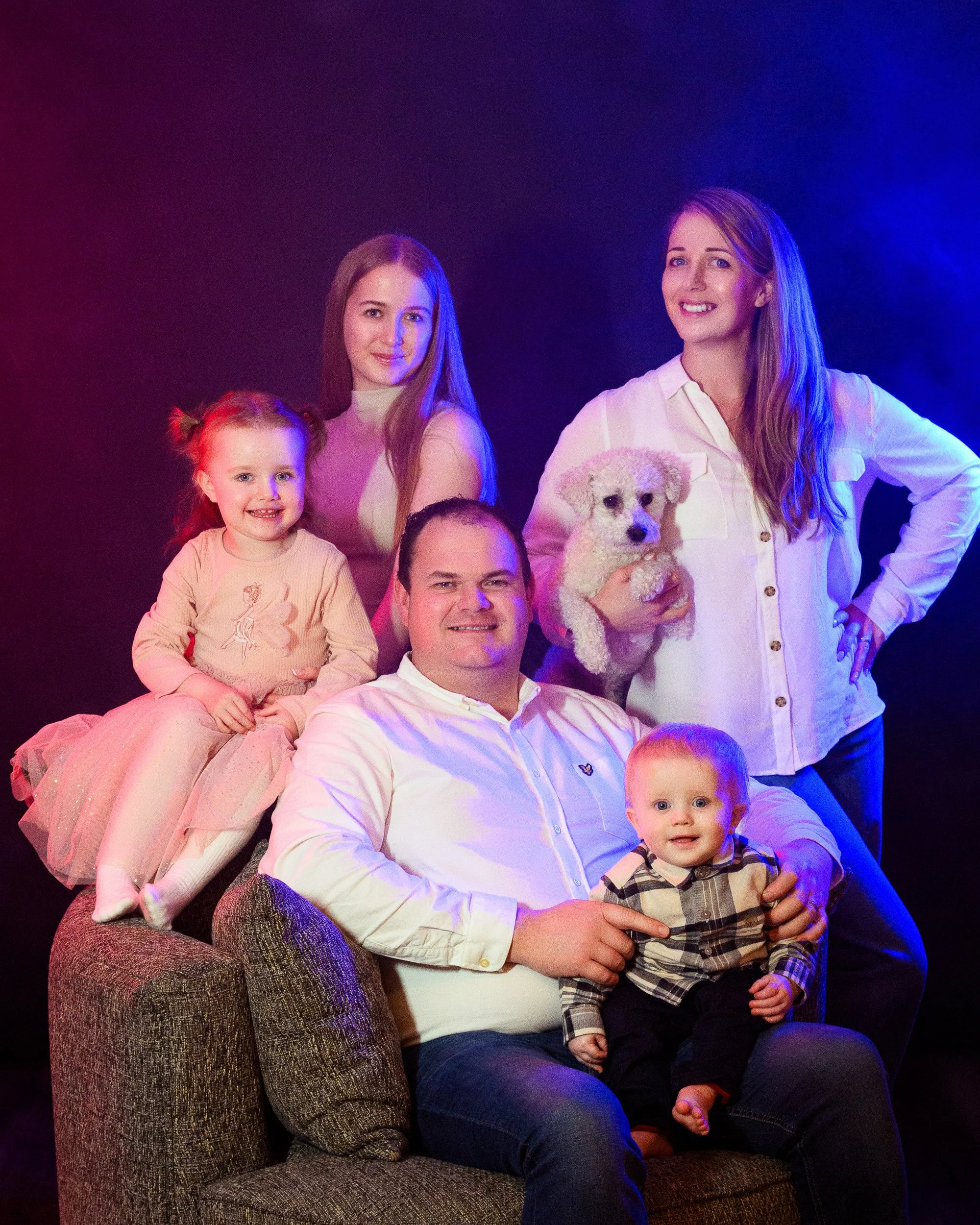 A large family group portrait with two women, two men, two children, and a dog, set against a dark background with subtle coloured lighting.
