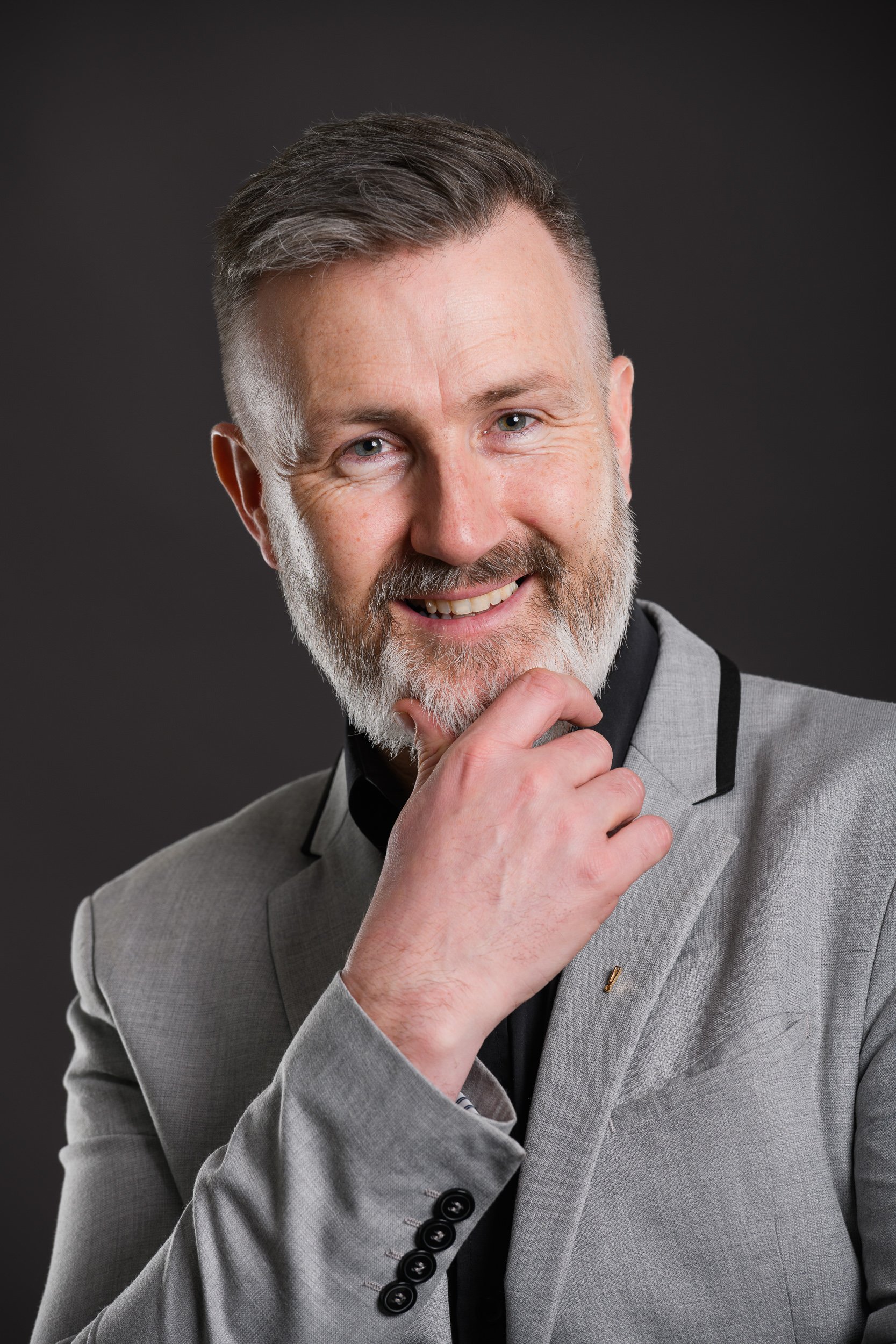 Professional portrait of a middle-aged man with grey hair and beard, wearing a suit and resting his chin on his hand, smiling against a dark background.
