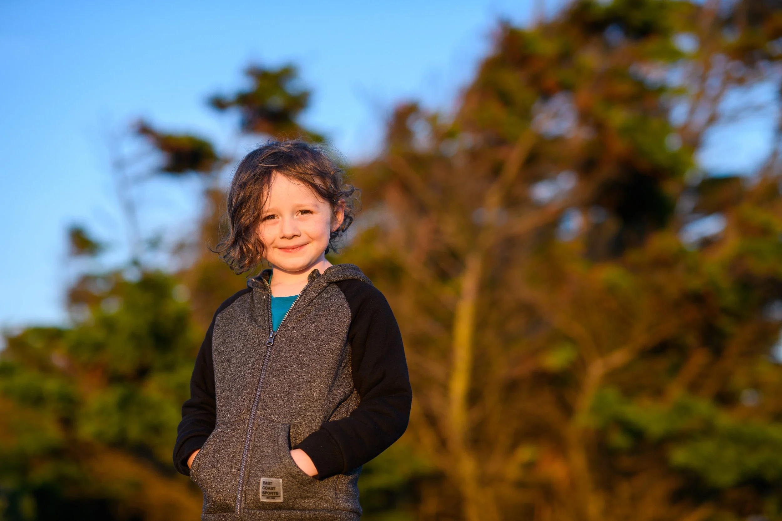 A young child with curly hair smiling outdoors during sunset with trees in the background.
