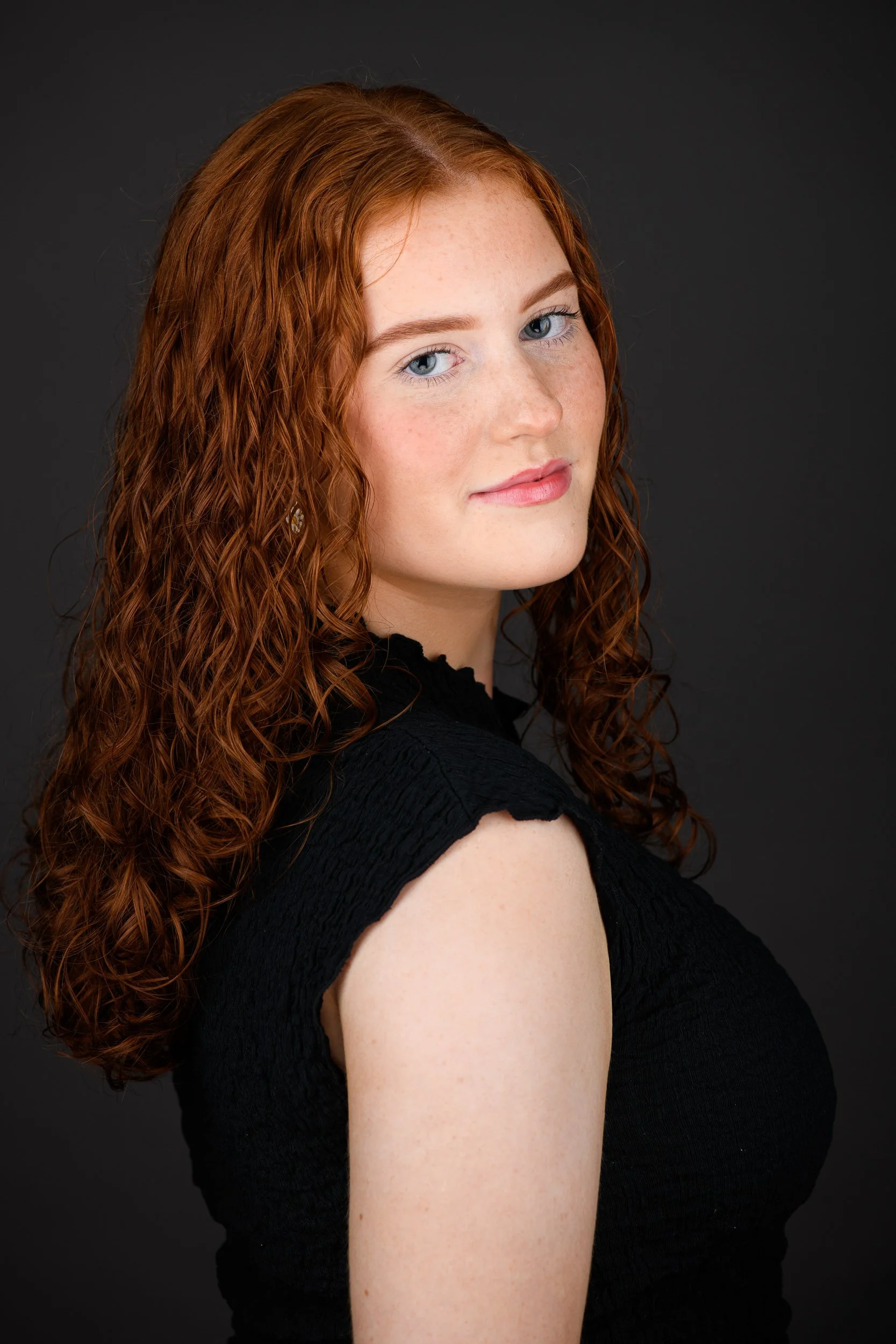 Portrait of a young woman with red curly hair, wearing a black sleeveless top, against a dark background.