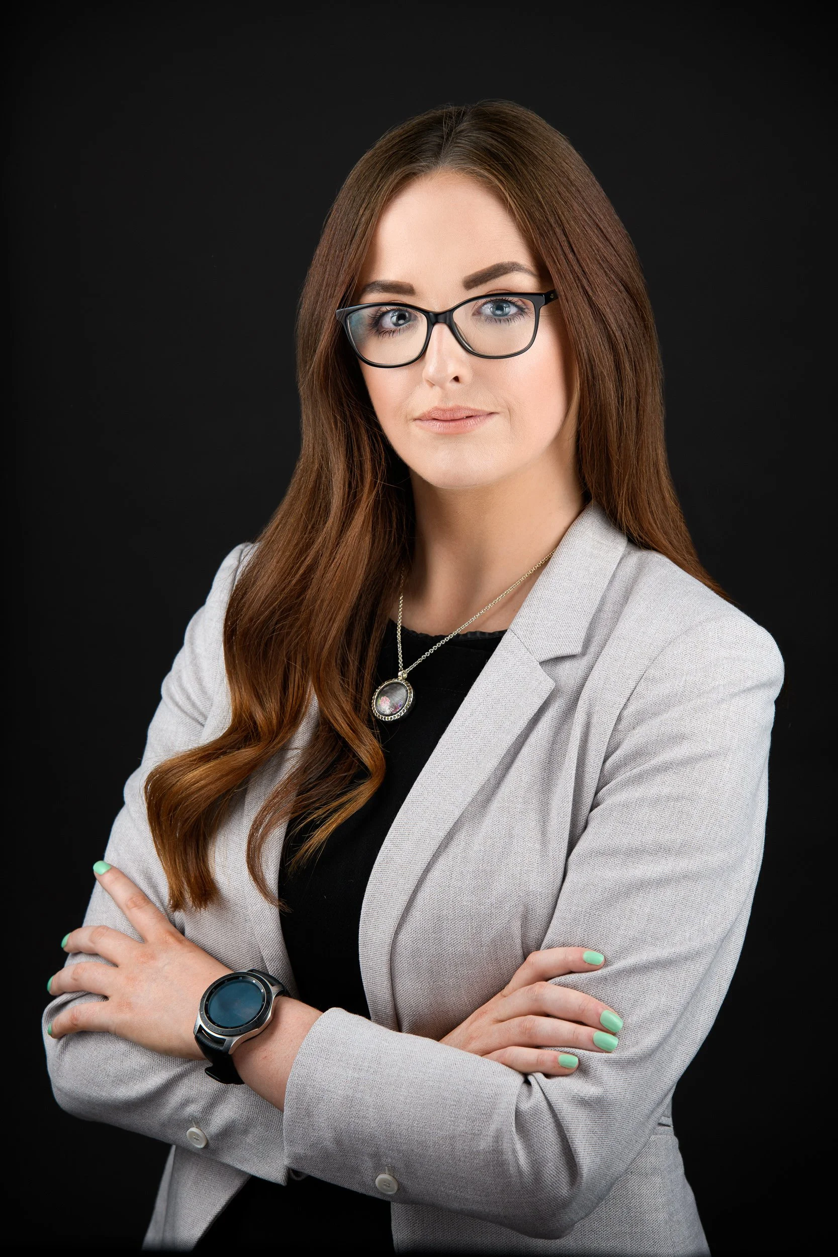 Professional portrait of a woman with auburn hair and glasses, wearing a light grey blazer over a black top, posed with arms crossed against a black background.

