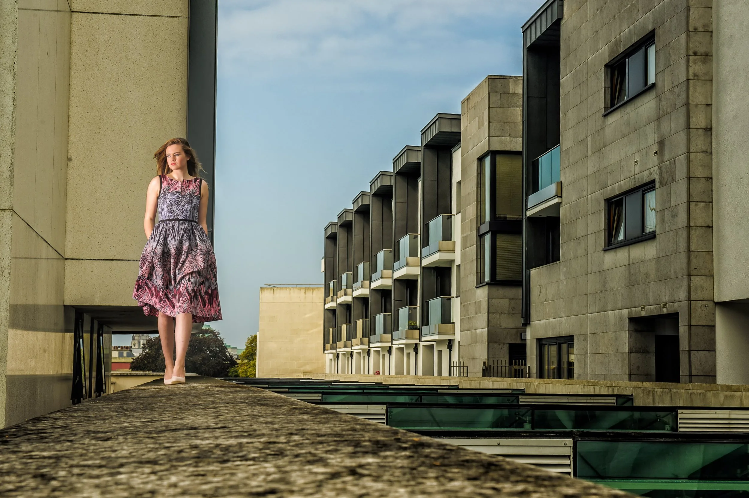 The Sheraton Hotel Athlone Fashion portrait for Burgees Athlone of a woman walking along a concrete ledge between modern architectural buildings
