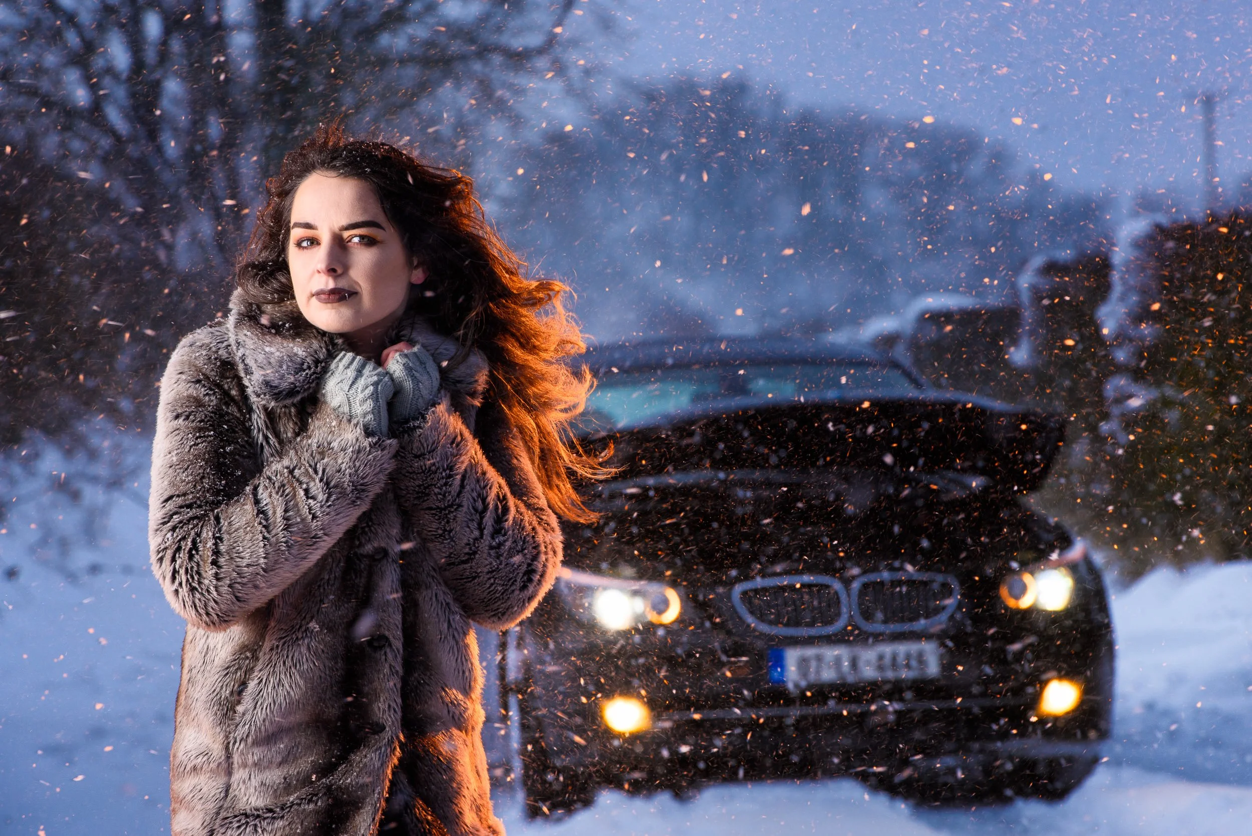 Cinematic winter portrait of a woman in a fur coat standing in snowfall with car lights behind