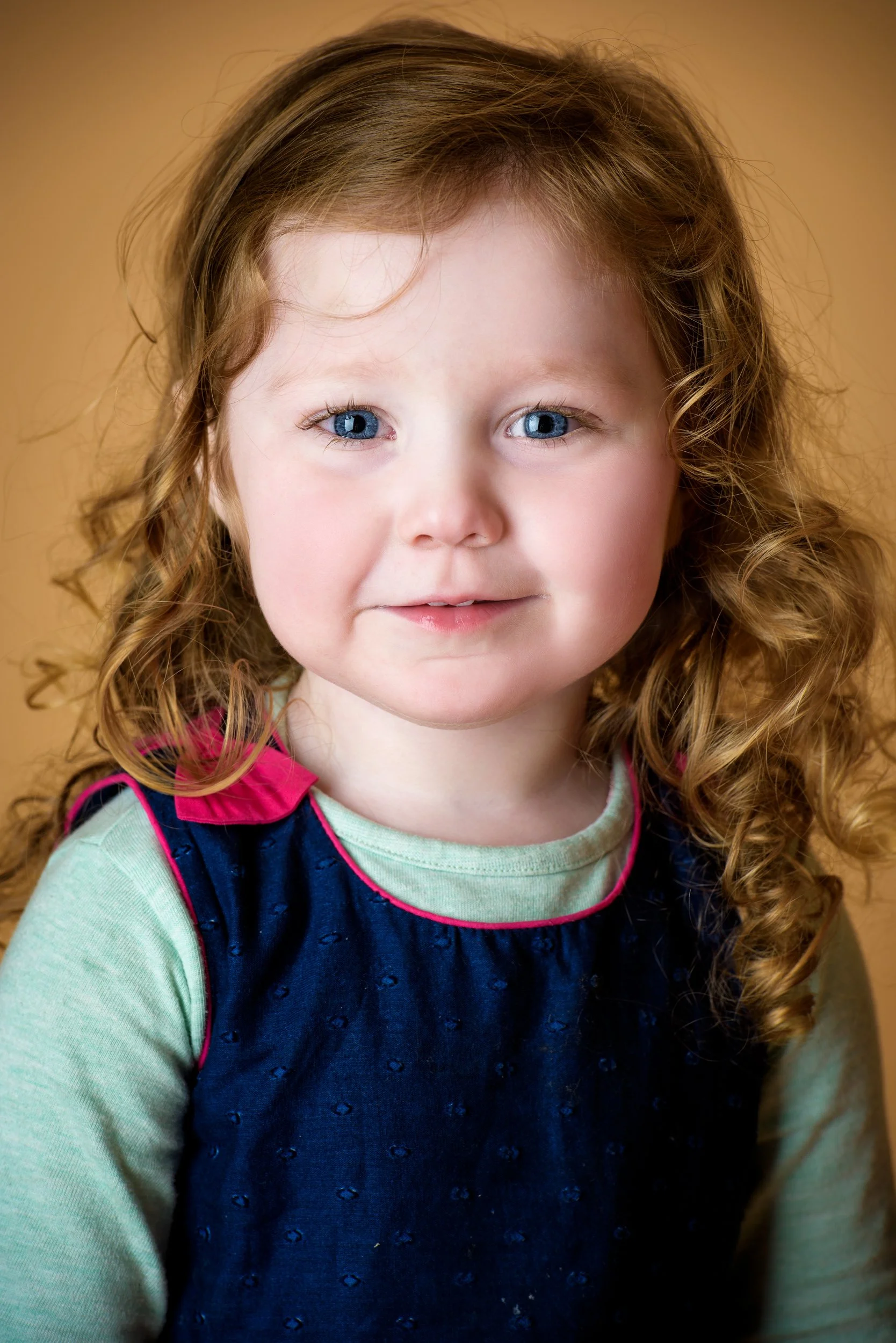 A young girl with curly red hair and blue eyes, wearing a green shirt and a navy blue jumper with pink trim, smiling softly against a warm brown background.