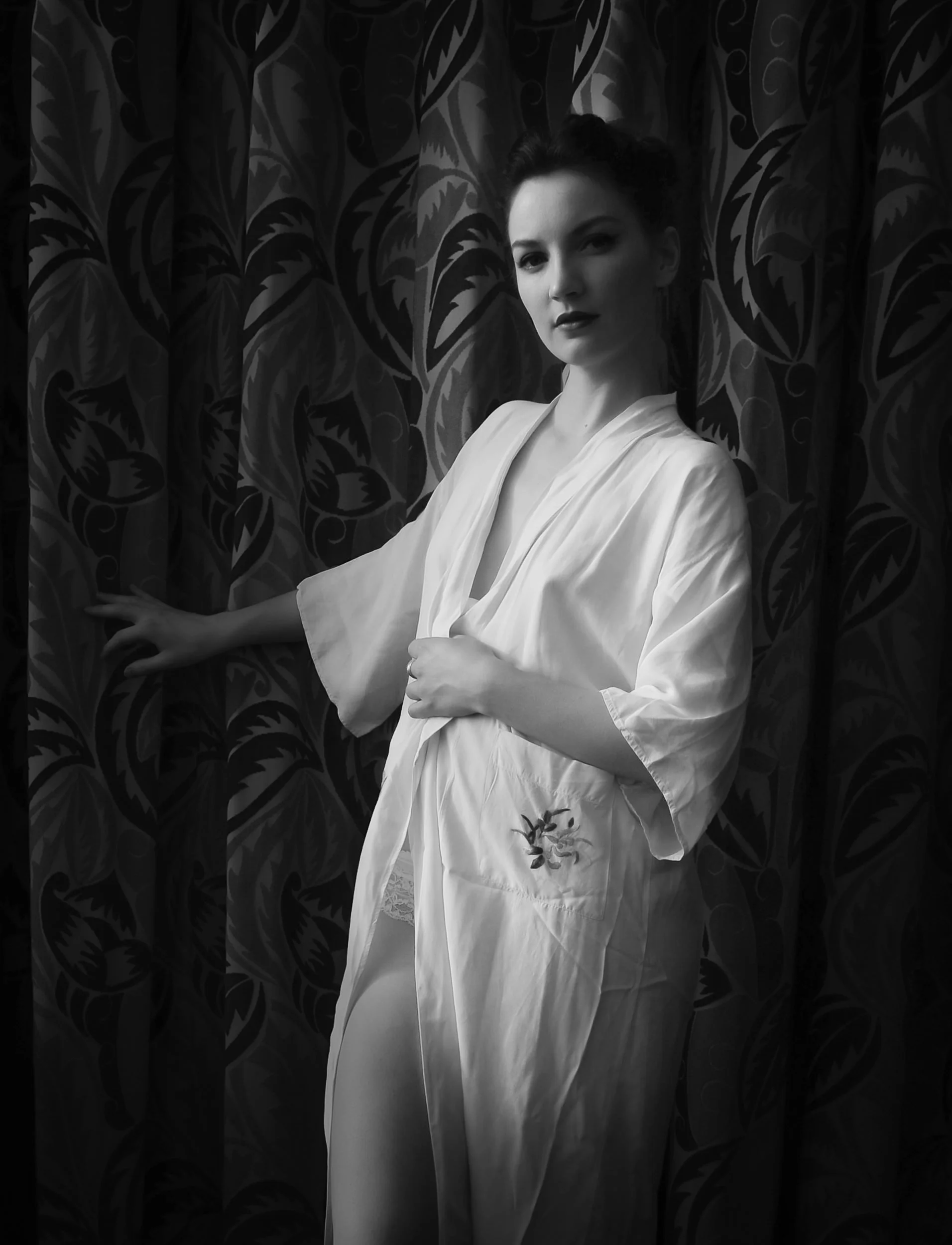 Black and white portrait of a woman in a silk robe standing against patterned curtains
