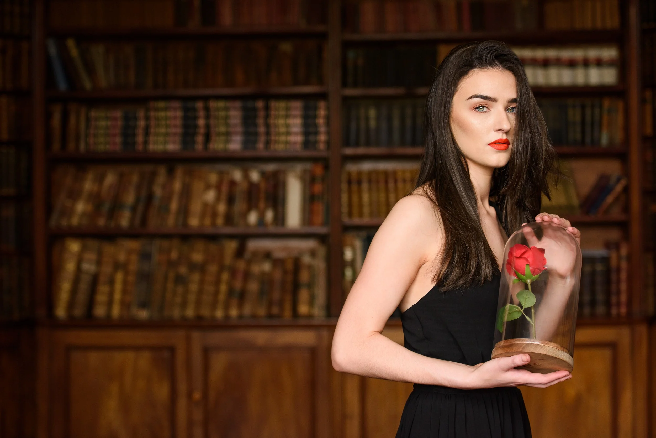 Conceptual portrait of a woman holding a glass dome with a red rose in front of a bookshelf

