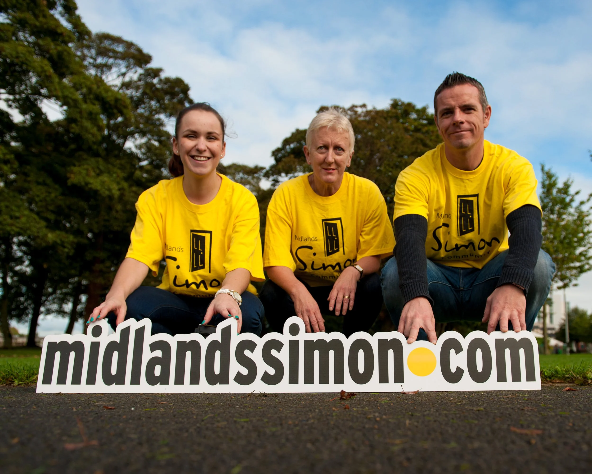 Three people wearing yellow t-shirts with 'Midlands Simon' printed on them, crouching on grass behind a sign that reads 'midlands-simon.com' during daytime with trees and a partly cloudy sky in the background.