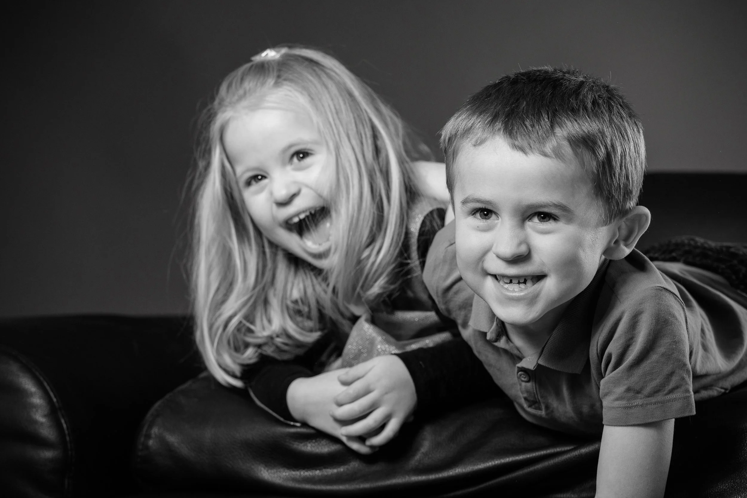Two children, a girl with long light hair and a boy with short dark hair, lying on a dark surface and smiling at the camera in black and white.