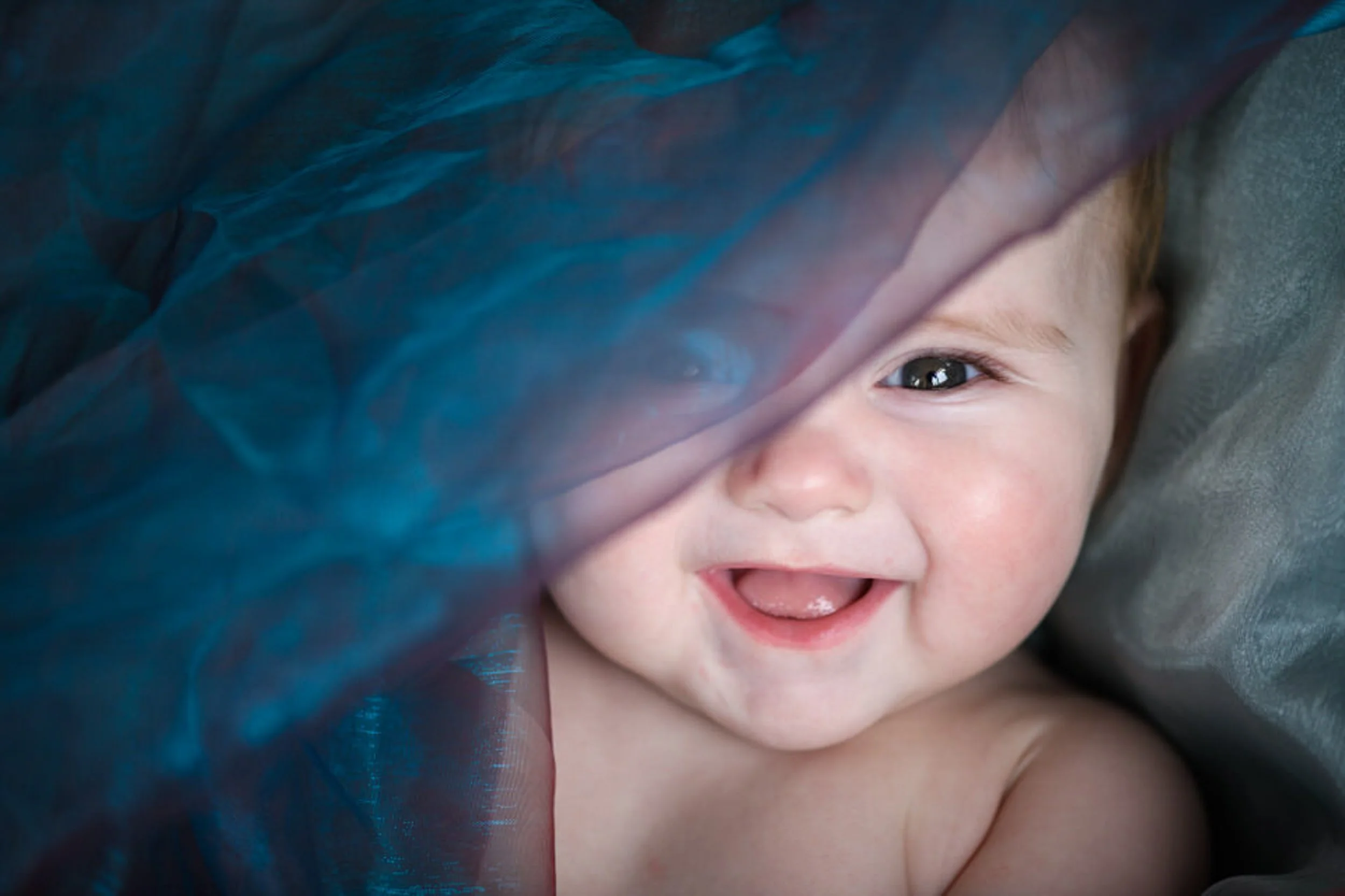 Smiling baby with blue eye partially covered by a black and blue fabric.