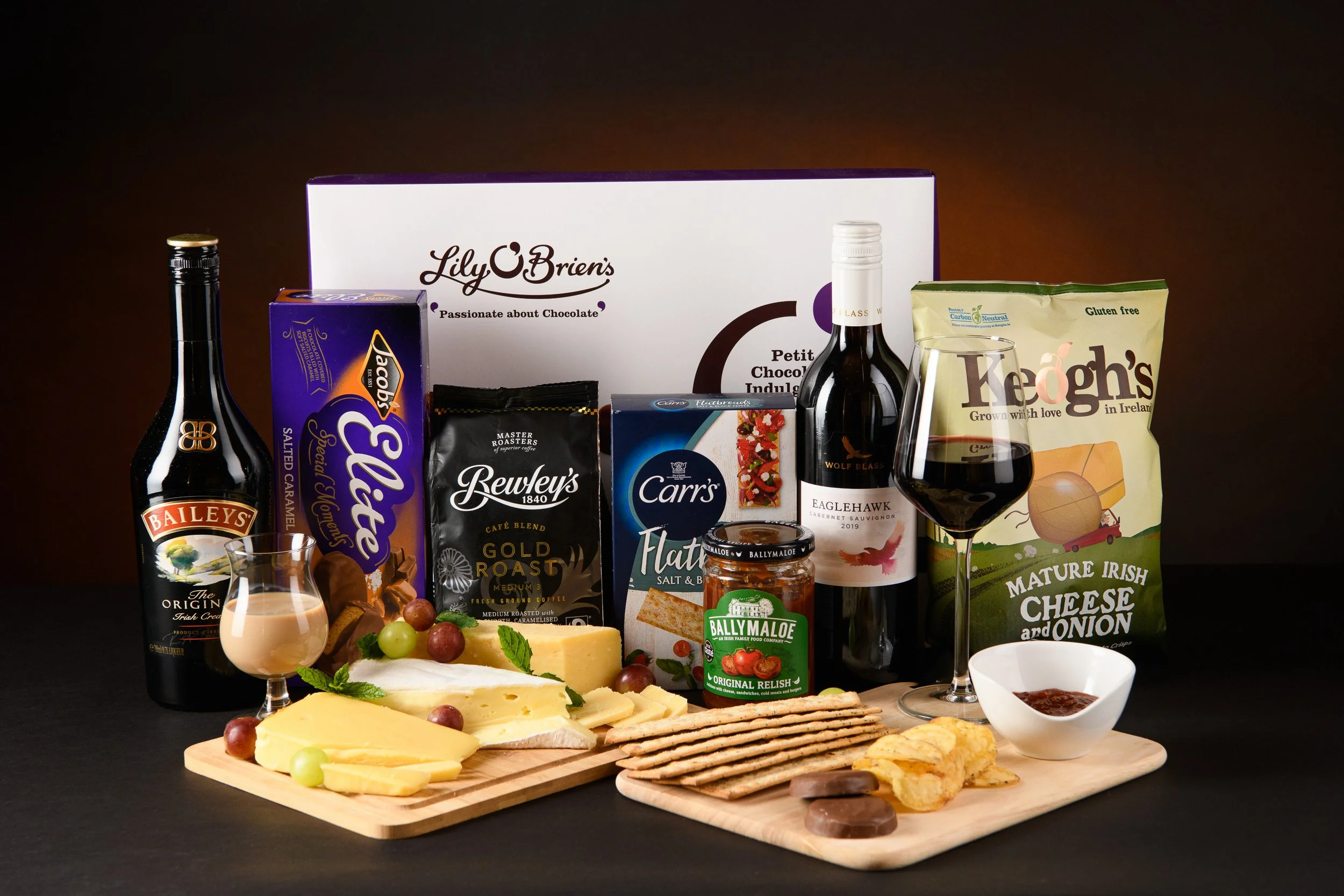 A food and drink gift basket includes cheese, crackers, grapes, a jar of relish, a bottle of red wine, a glass of red wine, a bottle of Bailey's Irish Cream, a box of chocolates, a bag of cheddar cheese and onion crisps, a bag of gluten-free Irish ch