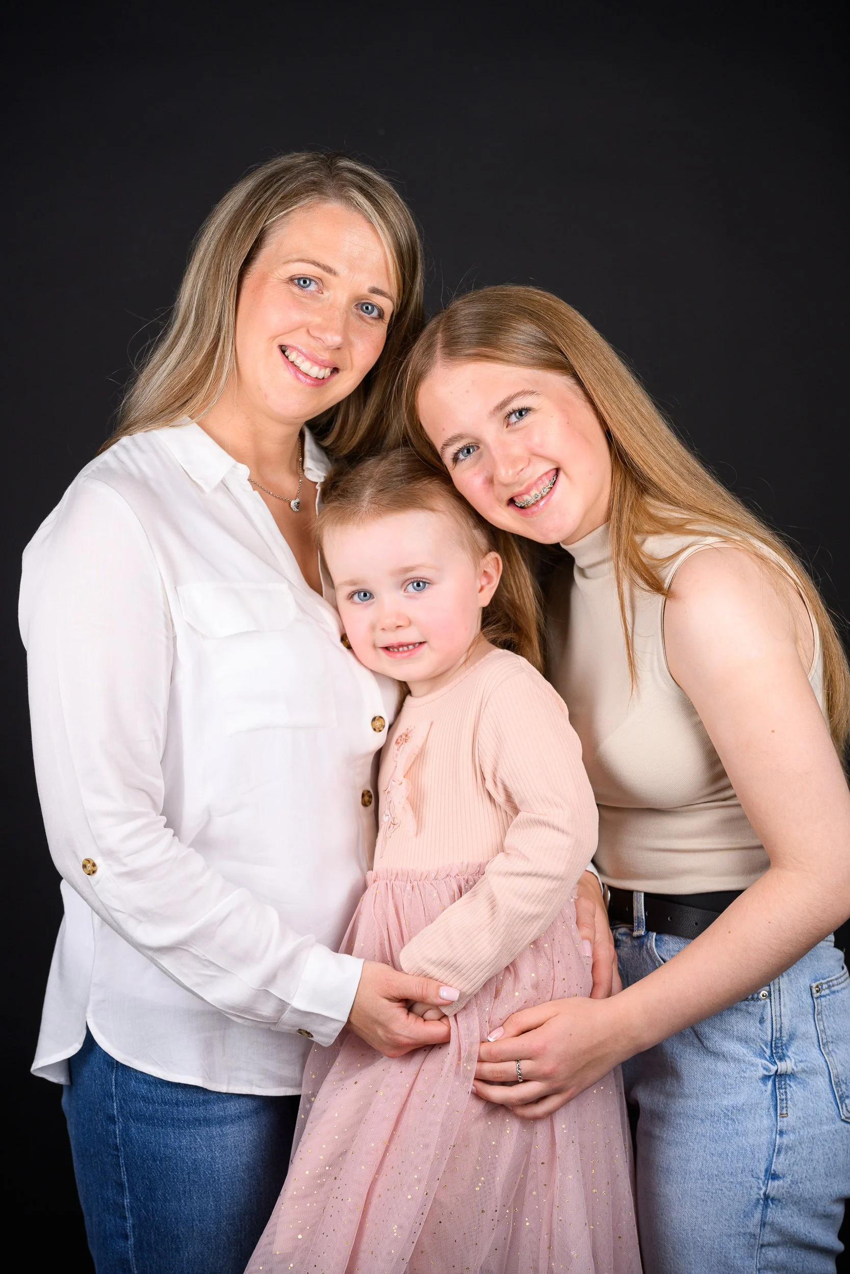 A mother with her two daughters smiling together against a black background.
