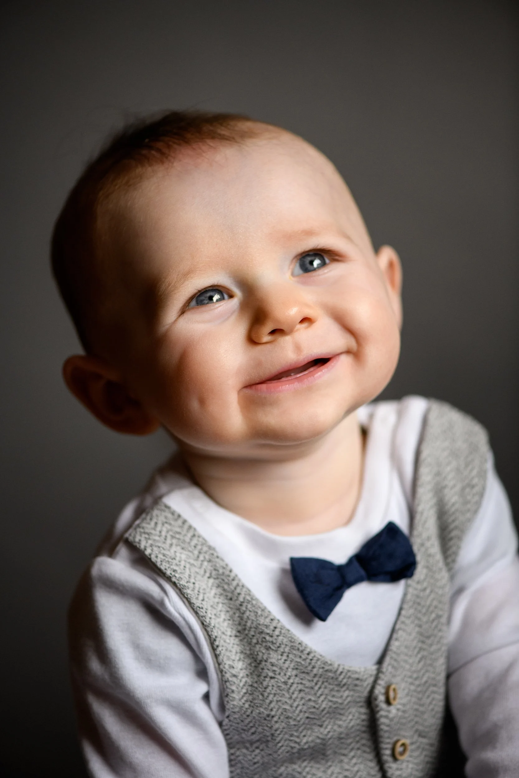 Ken Byrne Portrait Photography - A baby with blue eyes wearing a bow tie and vest, smiling against a dark background.
