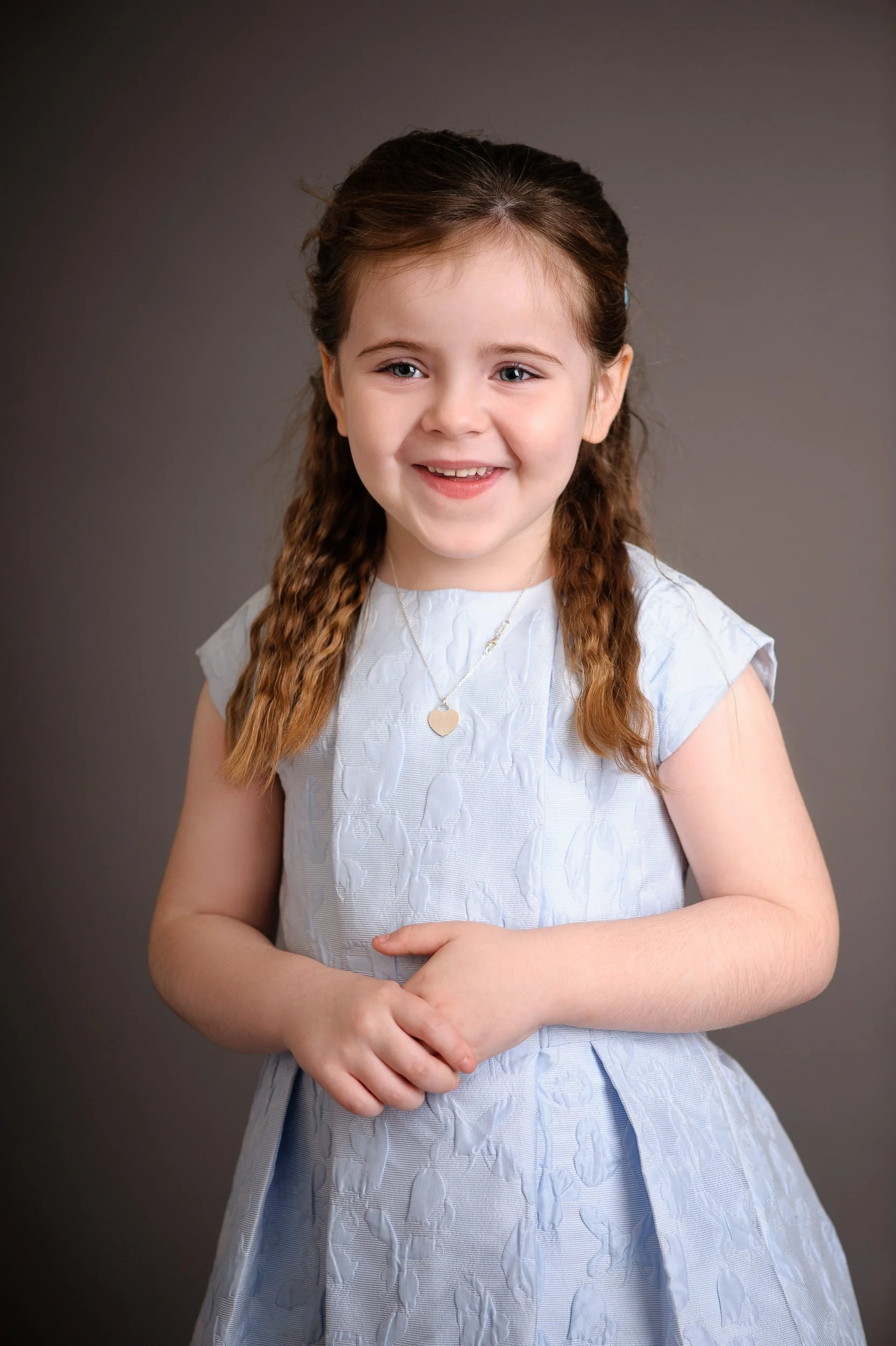 A young girl with brown hair smiling, wearing a light blue dress with textured pattern and a silver necklace with a heart pendant, standing against a gray background.