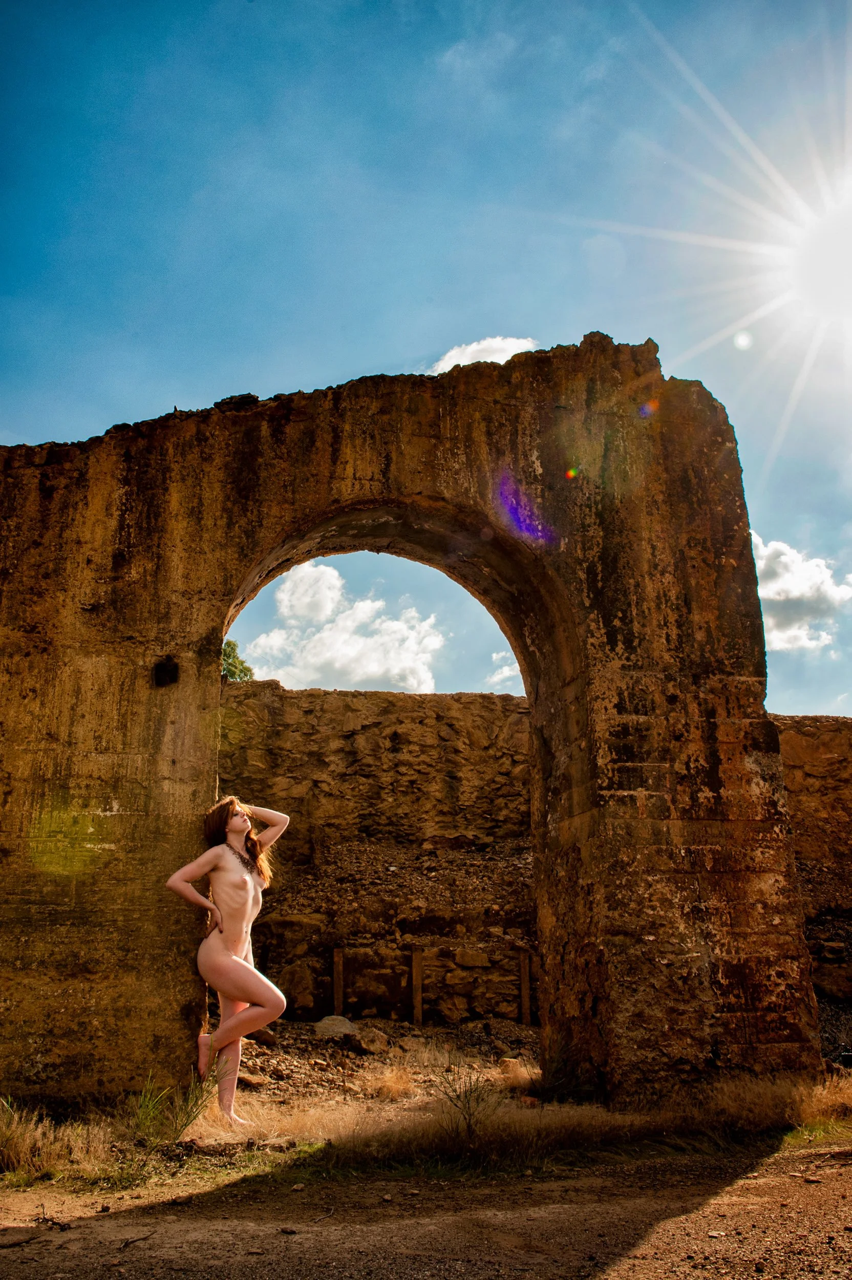 A woman with long red hair standing within a stone archway outdoors, framed by natural light and sky.
