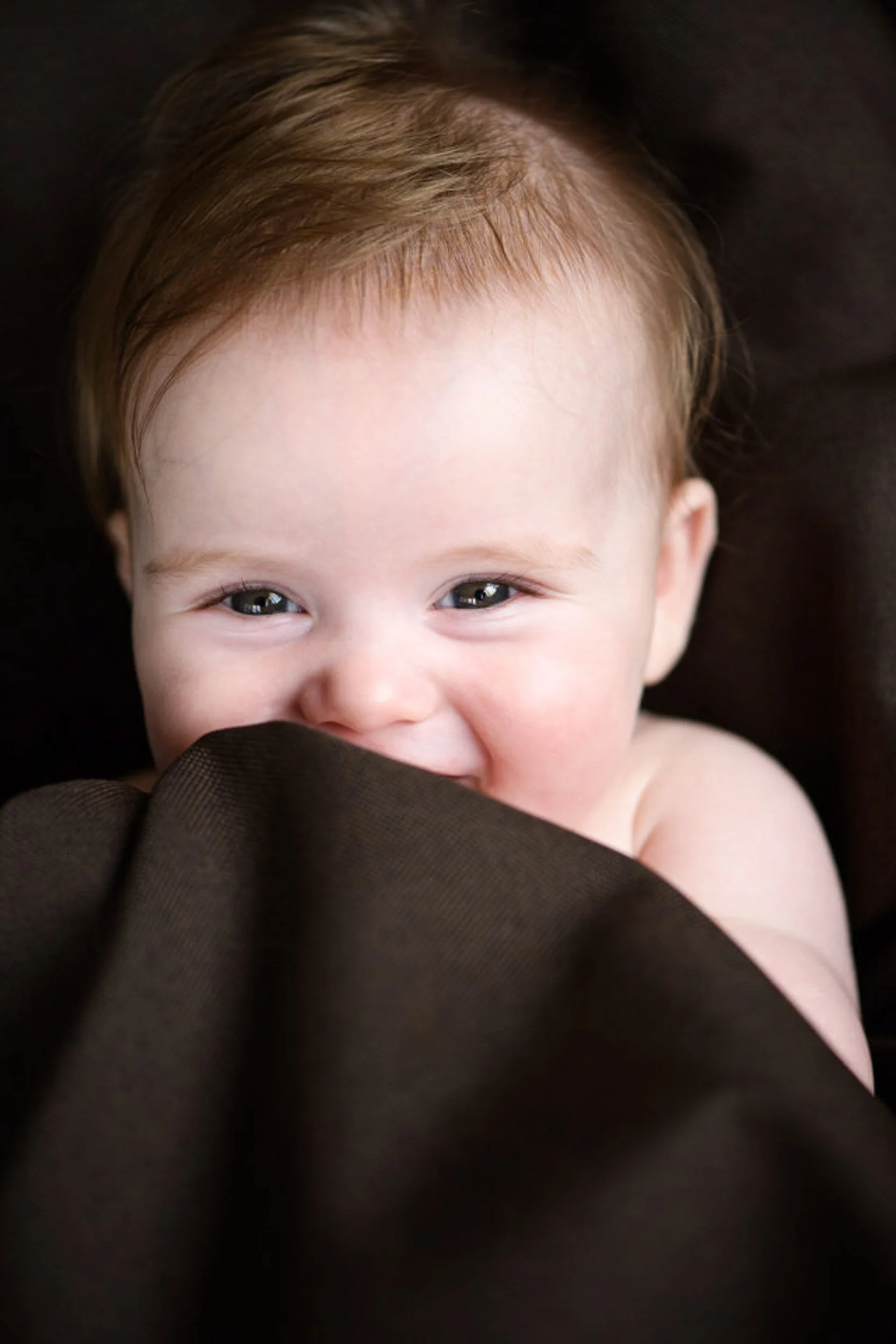 Close-up of a smiling baby with light brown hair, partially covered by a dark brown cloth.