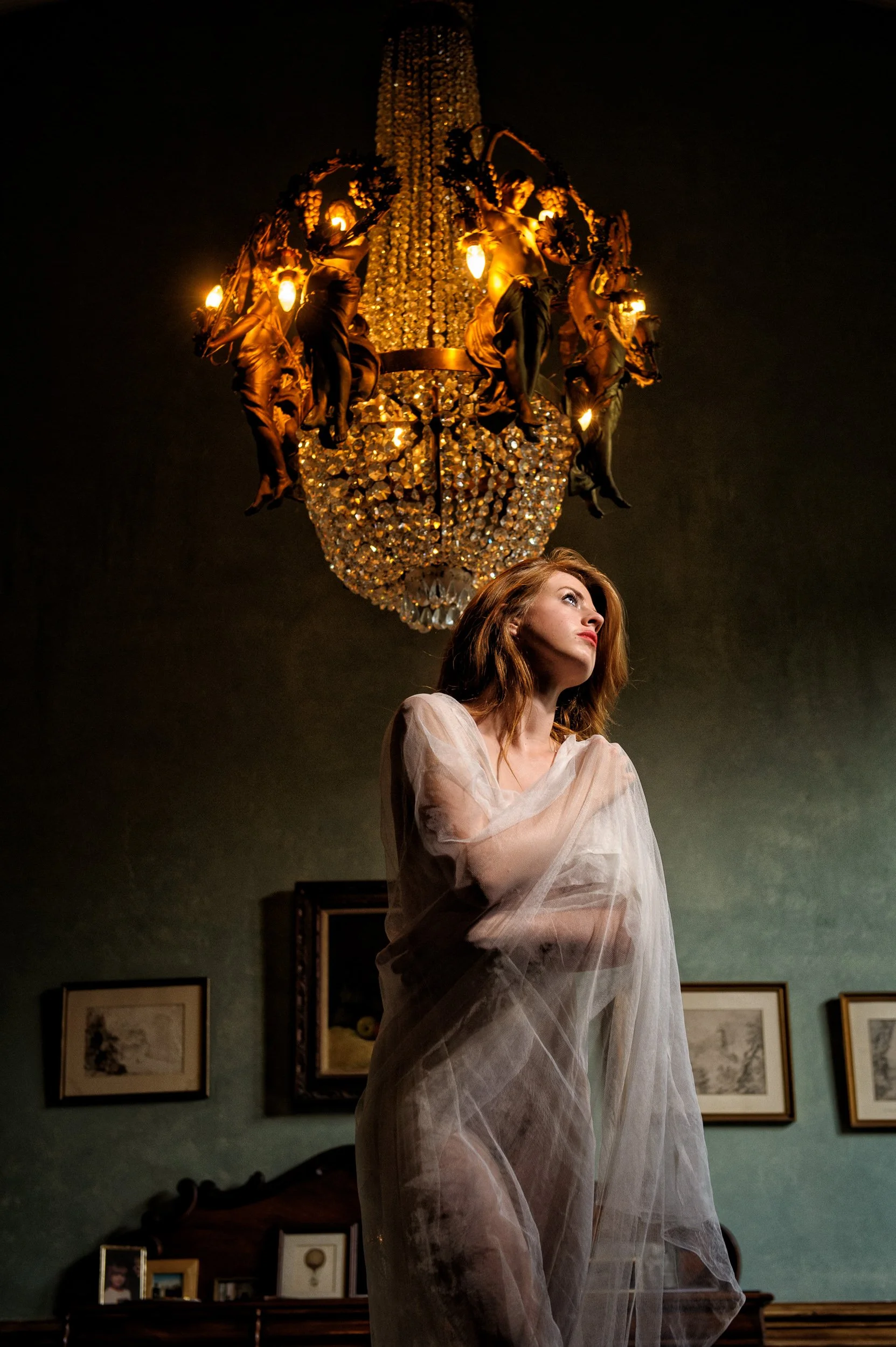 A woman with red hair wearing a sheer white dress, standing beneath an ornate chandelier in a richly decorated interior of Charleville Castle Ofally
