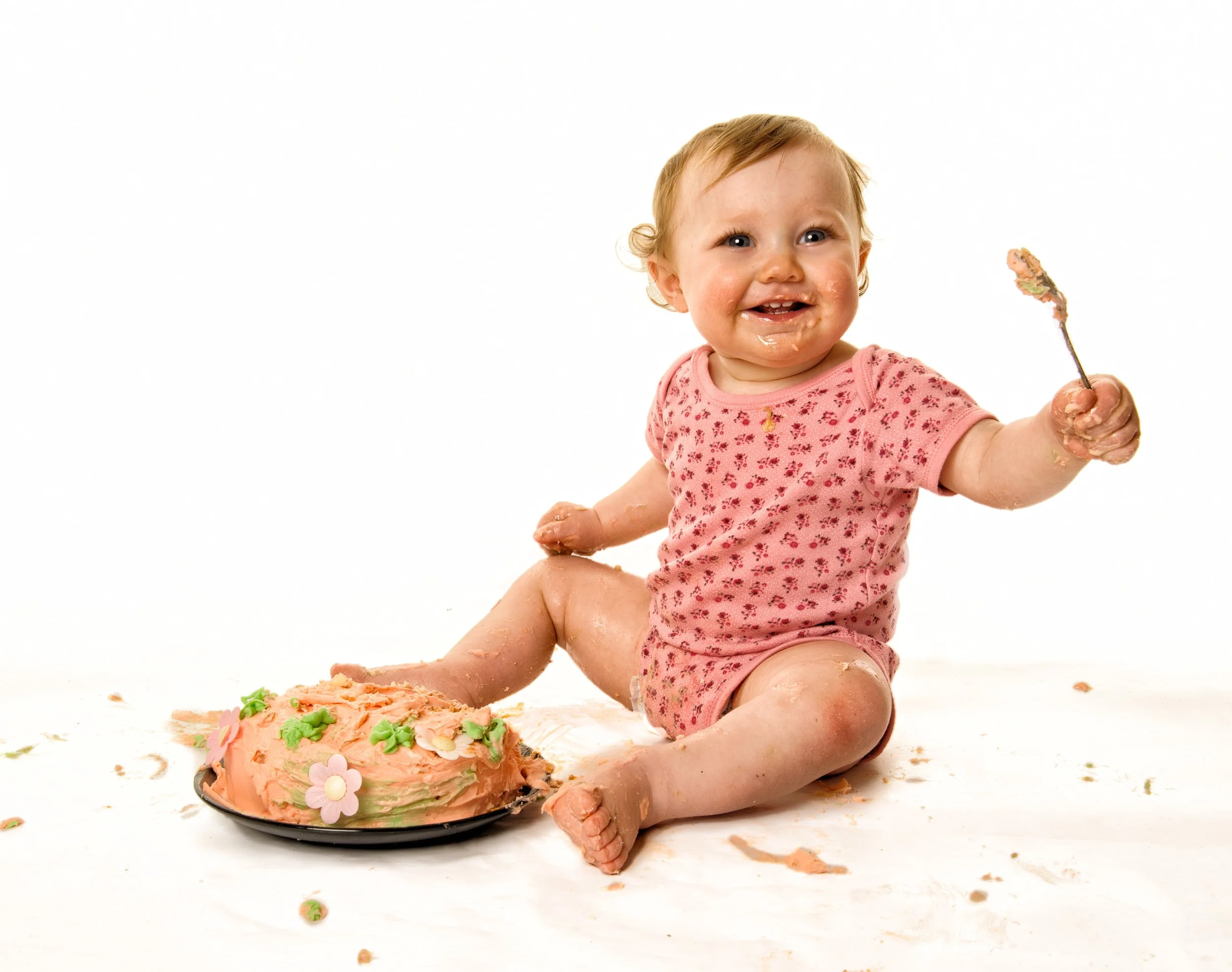 A baby with messy face and hands sitting on a white surface, holding a spatula, with a partially eaten pink and green decorated cake nearby.