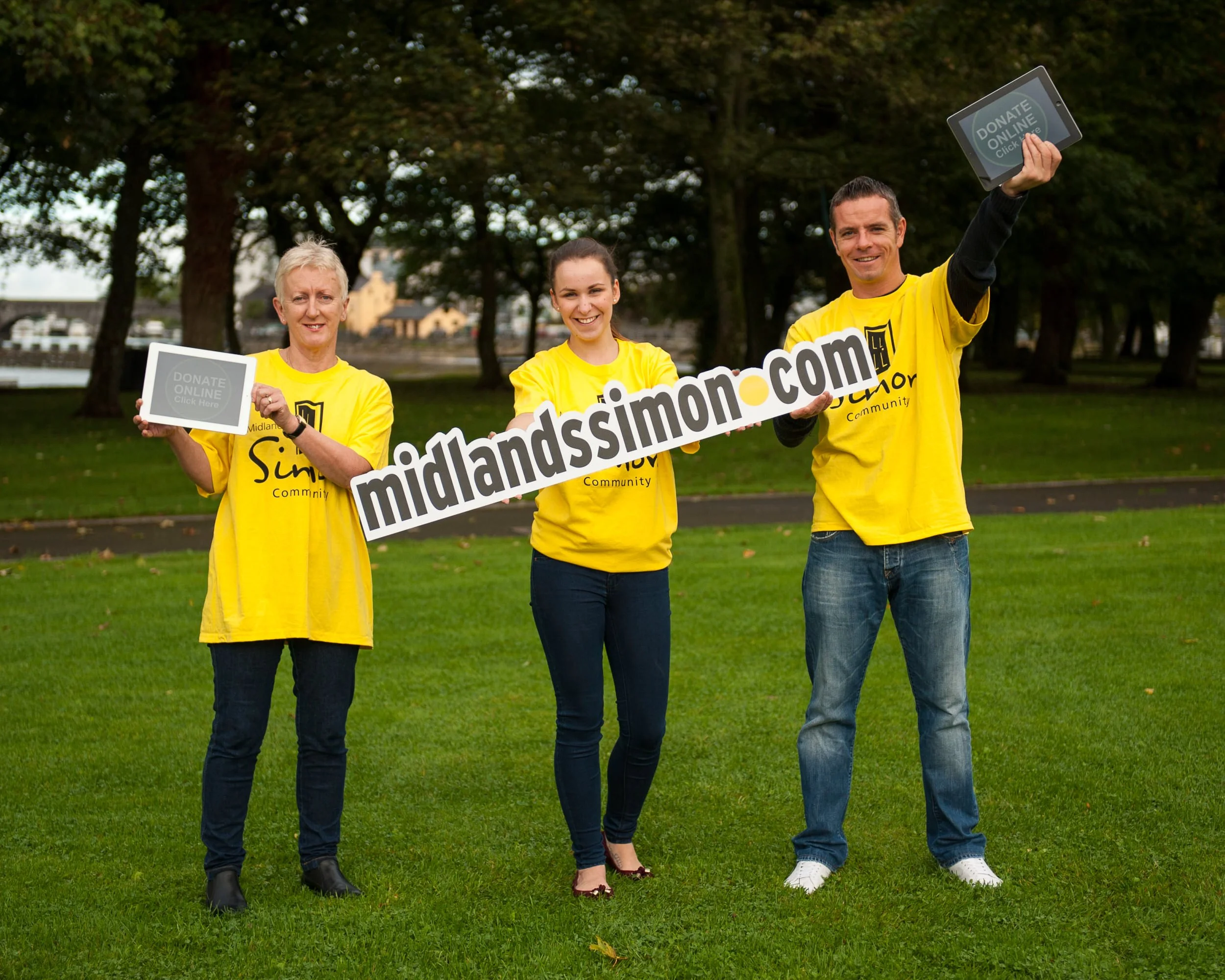 Three volunteers in yellow shirts holding a large MidlandSimmons.com sign and donation tablets while standing on a grassy park with trees.