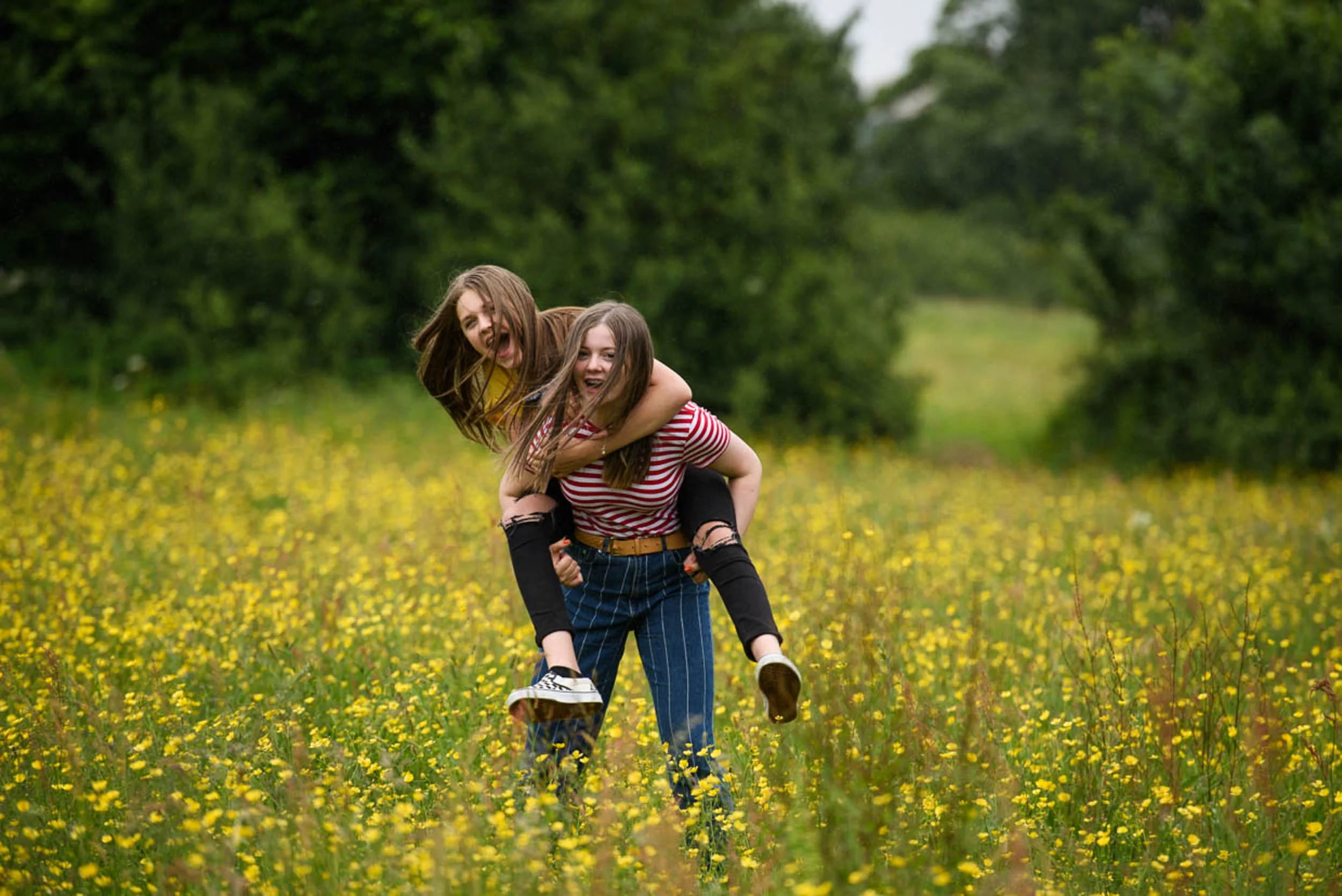 Two young women enjoying a playful piggyback ride through a field of yellow flowers, surrounded by green trees.