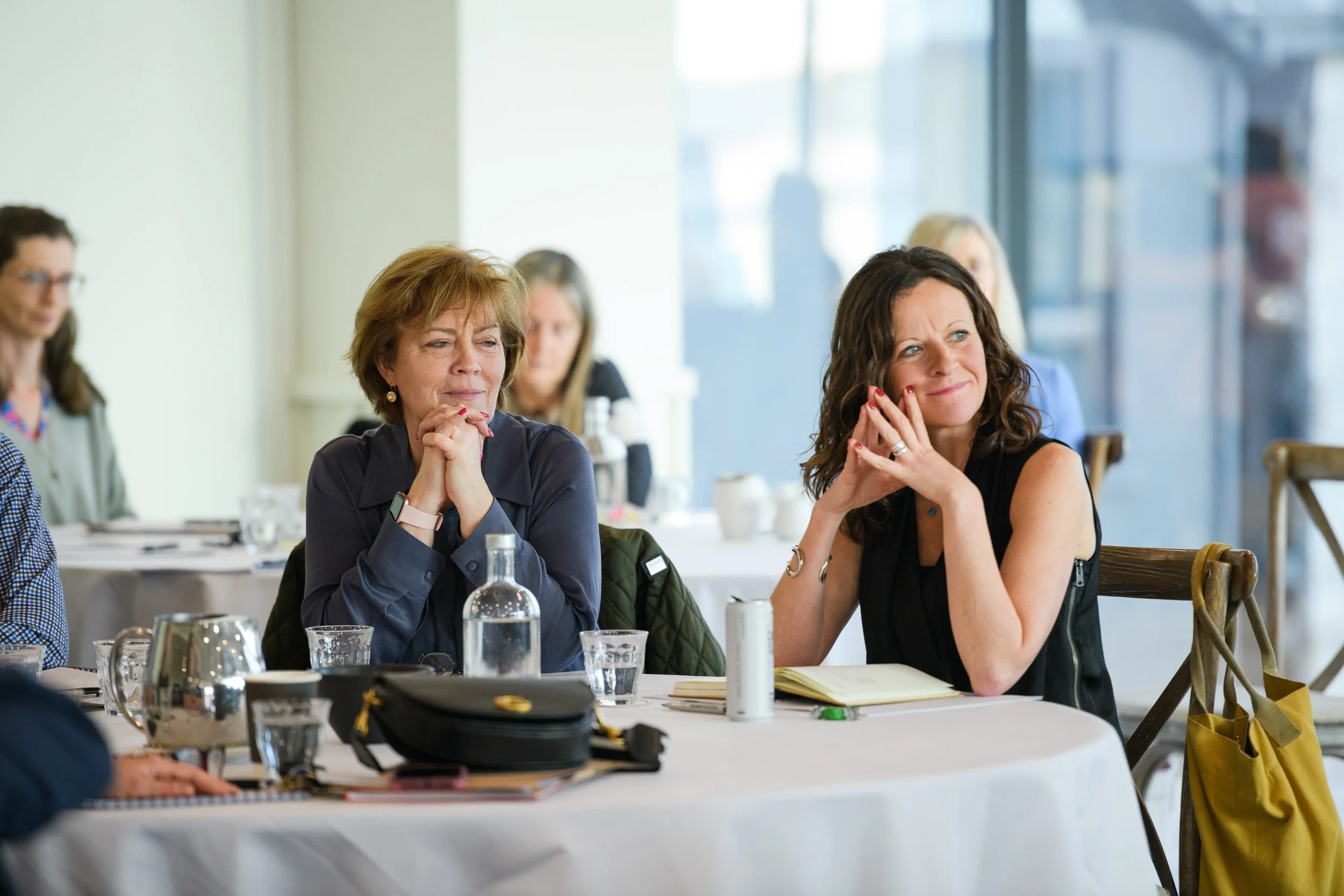 Women attentively listening during a meeting in a well-lit conference room with notebooks and glasses on the table.