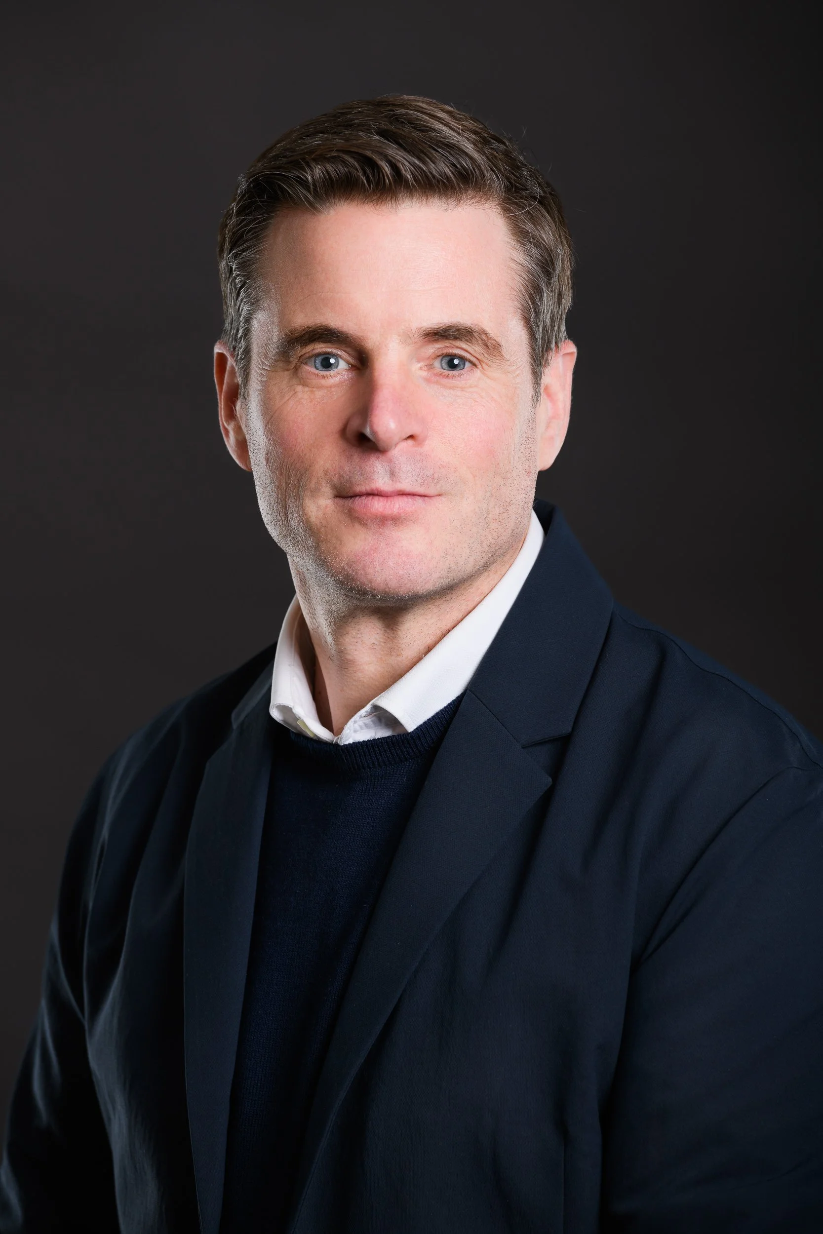 Professional portrait of a man with short brown hair and blue eyes wearing a blazer layered over a sweater and shirt, against a dark background.
