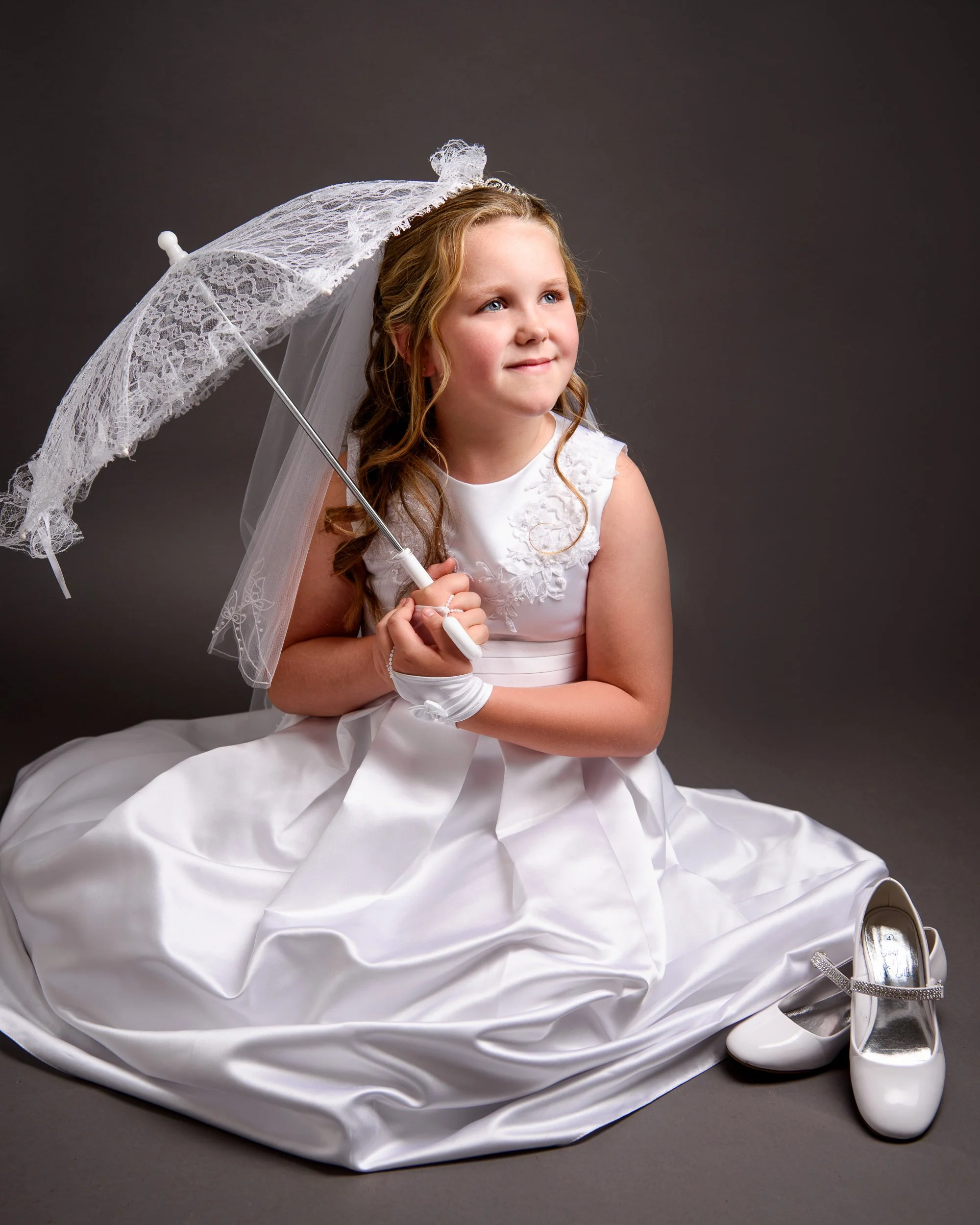 A young girl dressed in a white dress with a satin finish, holding a lace parasol, seated on a gray background. Two white shoes with bows are placed on the ground beside her.