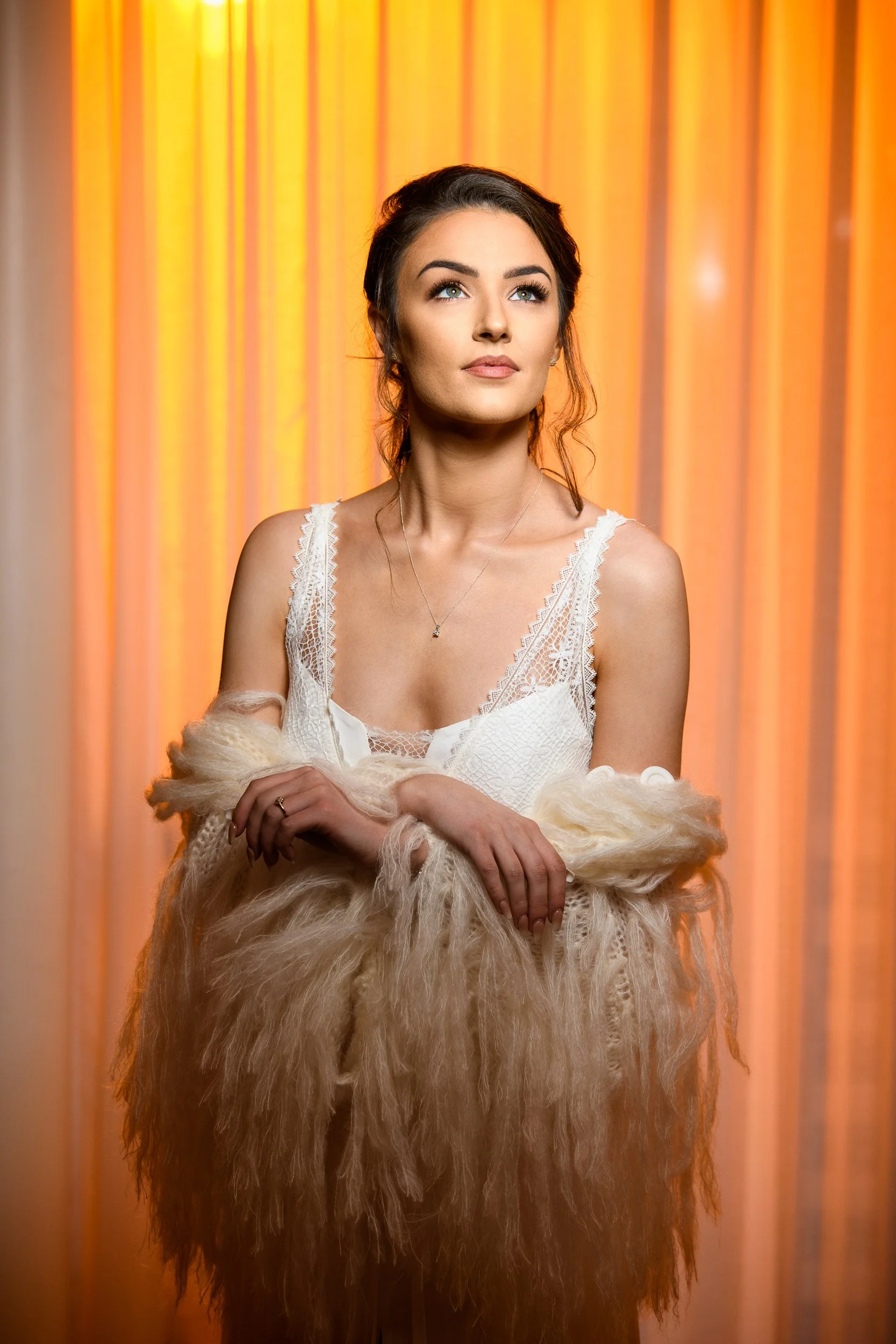 A Bride with dark hair styled in loose curls, wearing a white lace dress with a plunging neckline, holding a beige fur shawl, standing in front of orange curtains Westmeath Ireland 