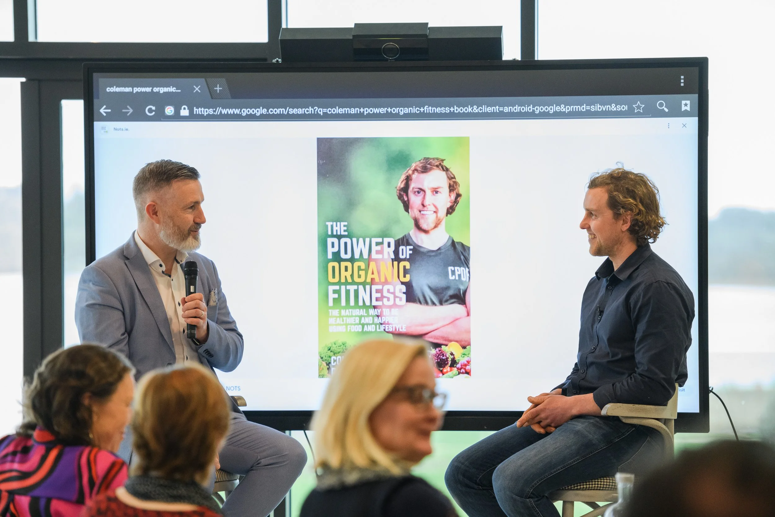 Two men are speaking in front of an audience, one holding a microphone. A large screen behind them displays a book cover titled "The Power of Organic Fitness." The audience comprises several women, some with glasses, in a well-lit room with floor-to-