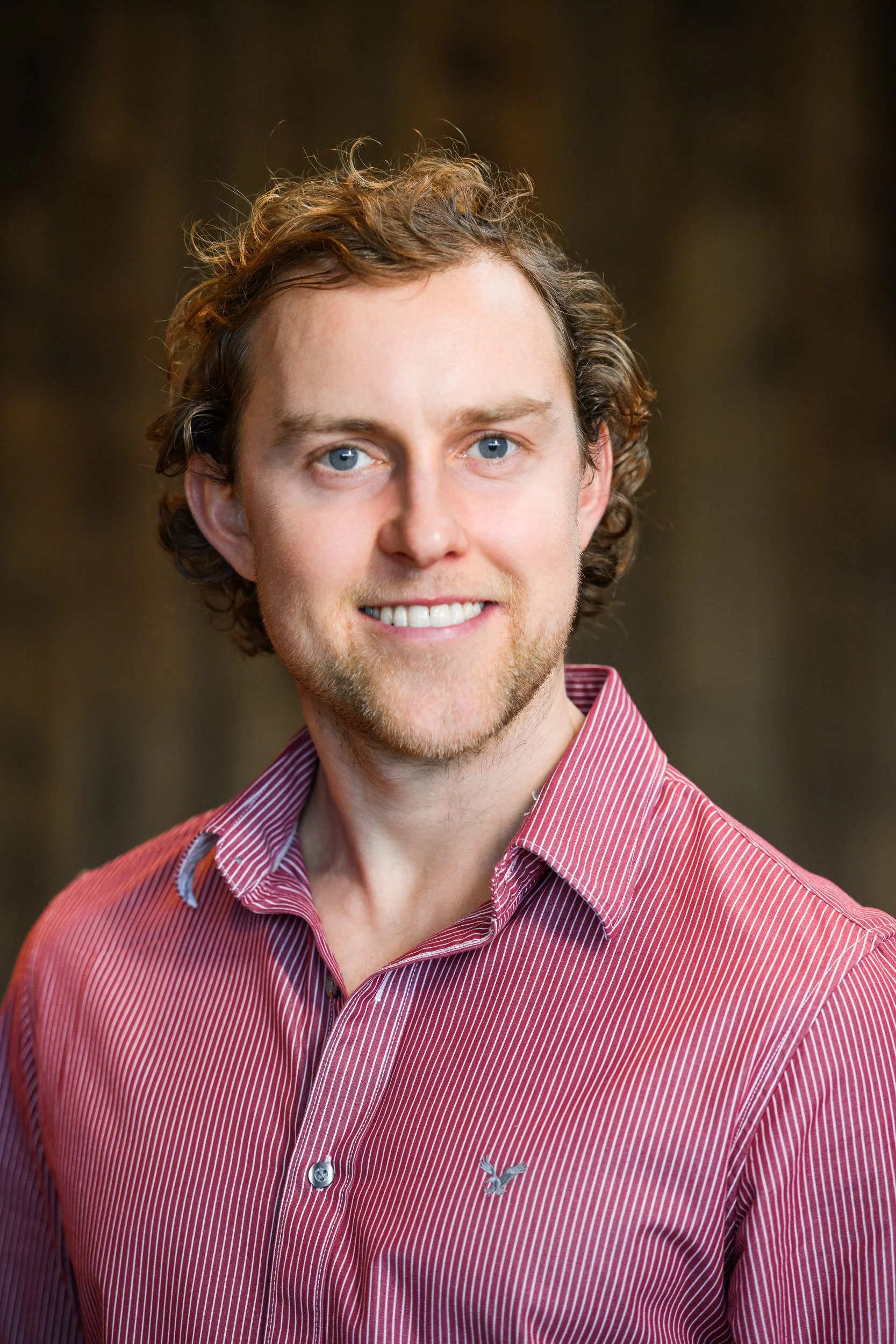 Portrait of a man with curly hair and beard wearing a striped shirt, smiling against a softly blurred dark background.
