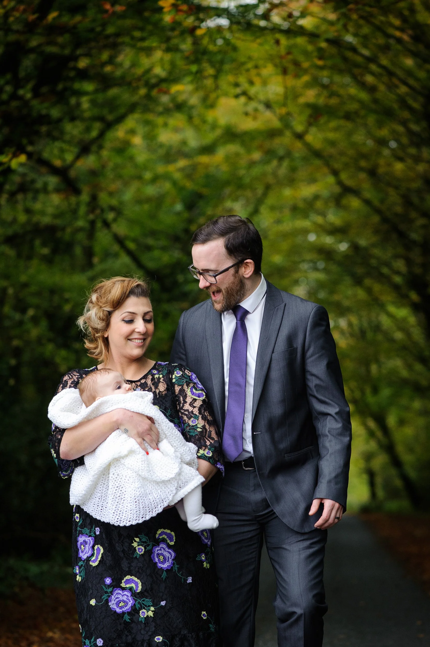 A man and woman holding a baby outdoors on a tree-lined path while smiling at a man in a suit, surrounded by greenery.
