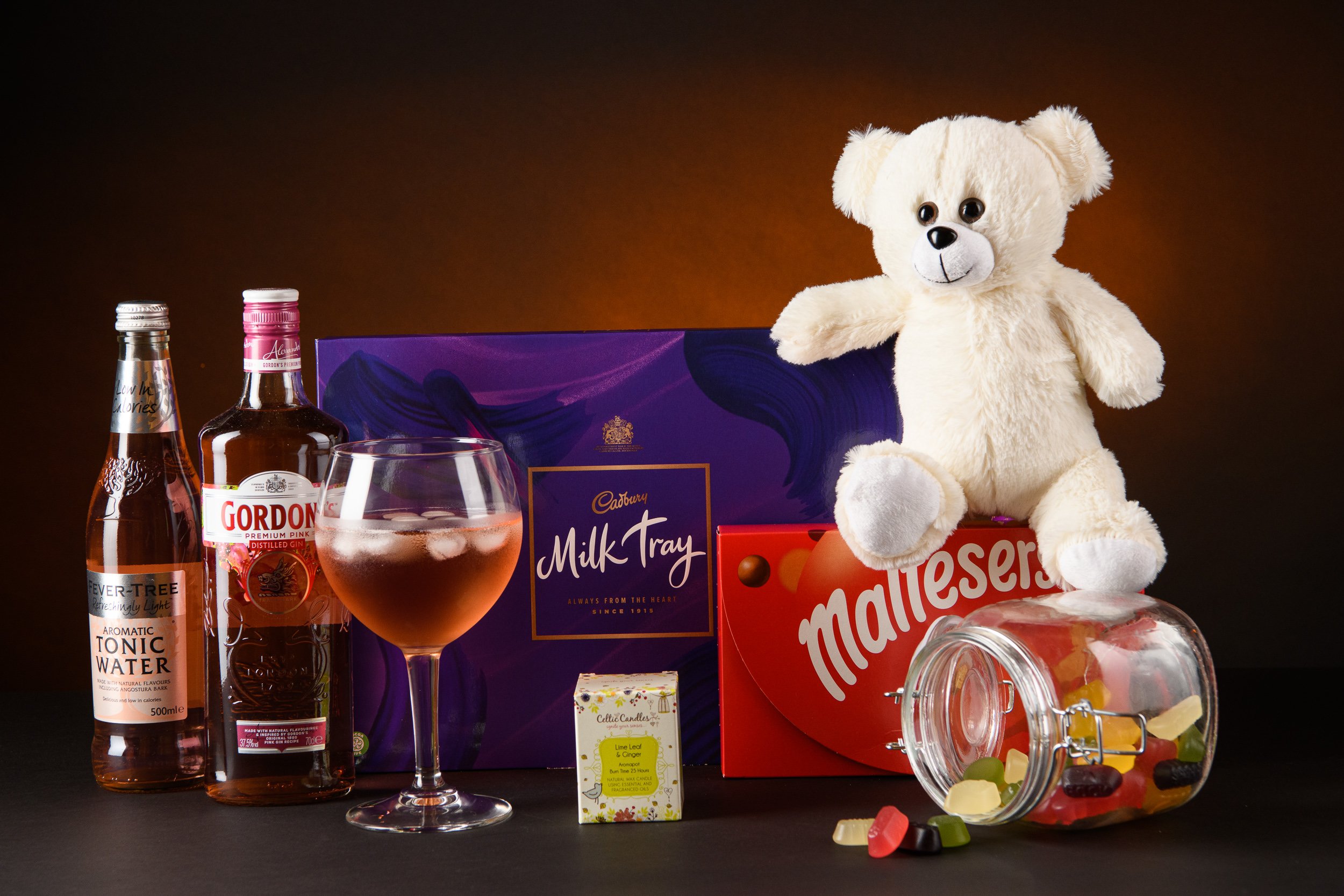 A styled product arrangement featuring confectionery, drinks, and a teddy bear, presented against a dark backdrop.
