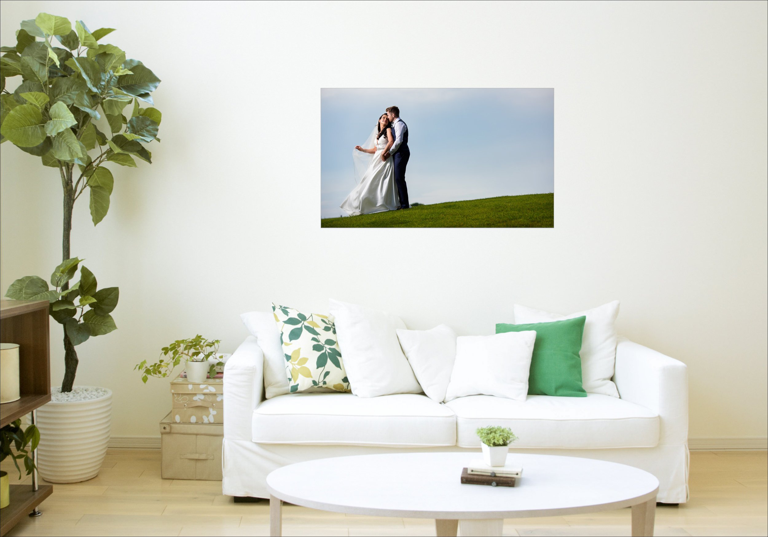 A white living room with a white sofa, decorated with green, white, and floral pillows. There is a small round white coffee table with a tiny potted plant and books, a large potted fig tree on the left, and a painting of a couple in wedding attire st
