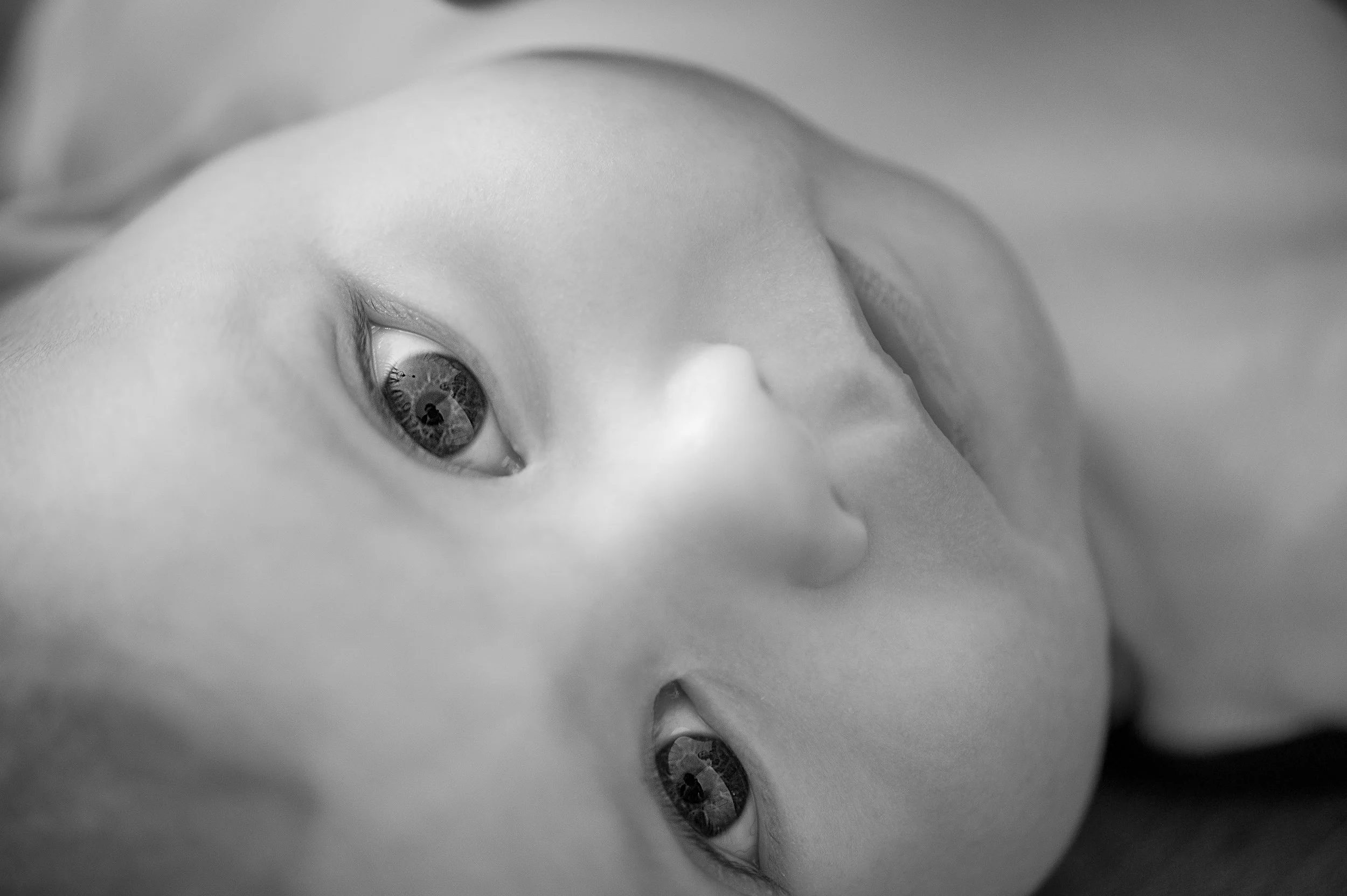 A close-up black and white photo of a newborn baby lying down, showing the baby's face and one eye with detailed eyelashes and iris.