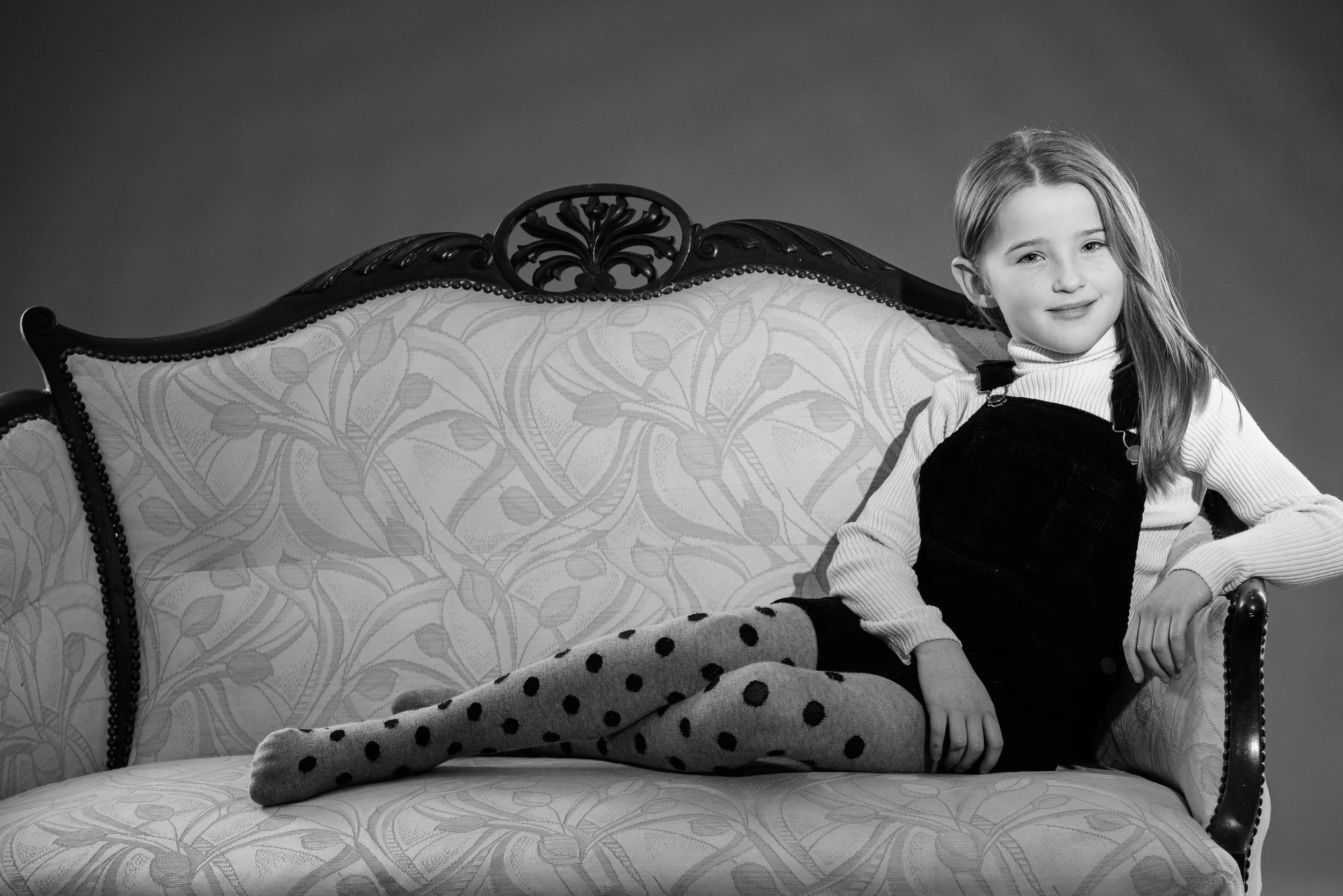 A young girl with long hair, sitting on a vintage-style couch with patterned upholstery, wearing polka dot tights, a turtleneck, and a jumper, smiling at the camera.