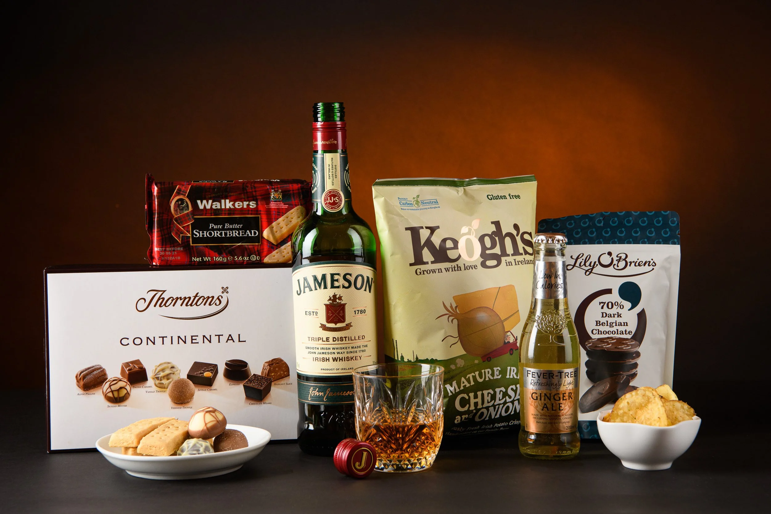 A curated selection of snacks and drinks including whiskey, chocolates, crisps, shortbread, and soft drinks styled together on a dark background.
