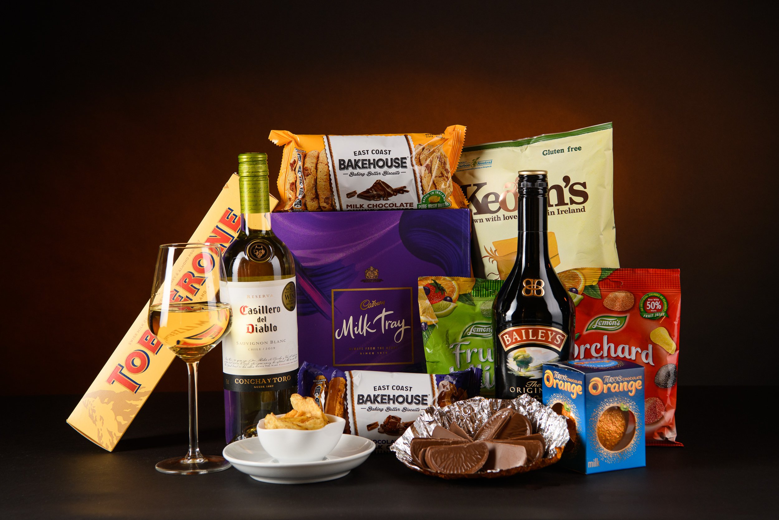 An assortment of snacks and beverages including wine, chocolates, crisps, biscuits, and confectionery arranged on a dark surface with a styled studio background.