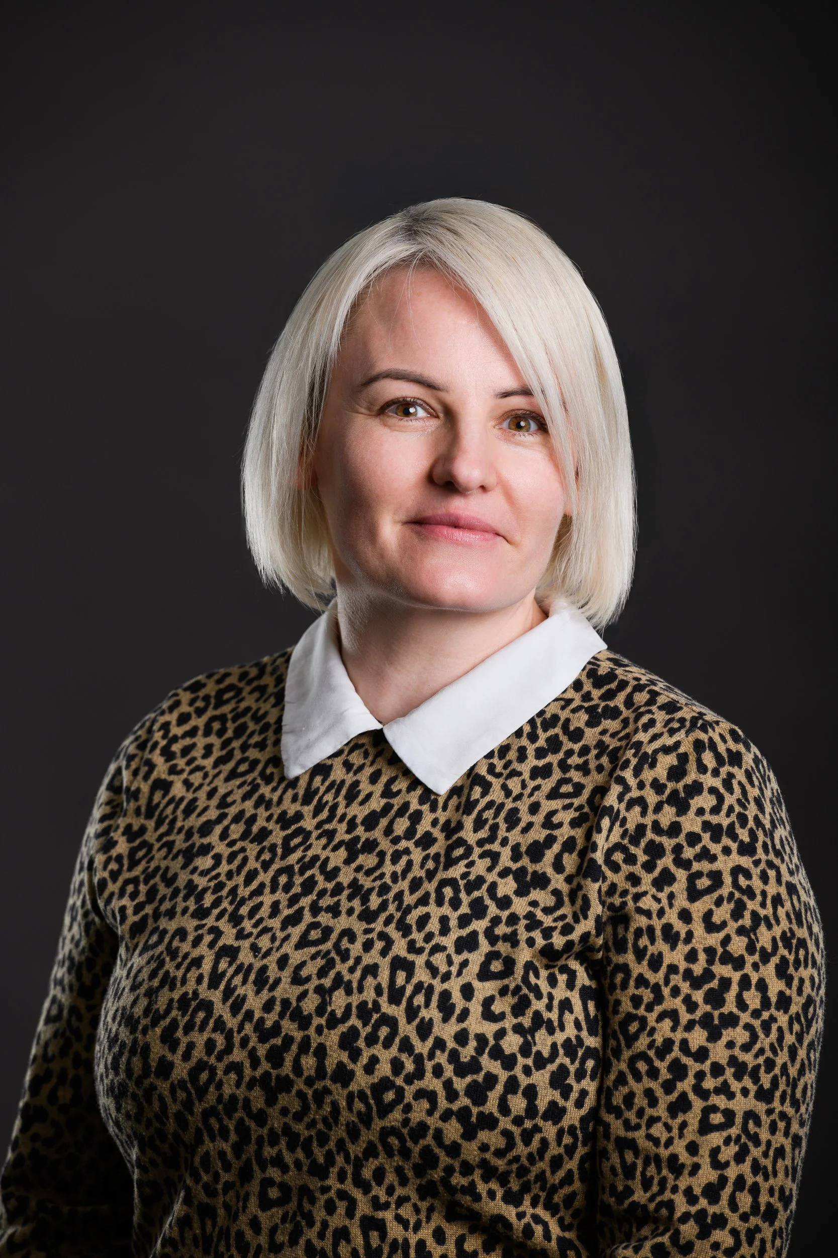 Professional portrait of a woman with short blonde hair wearing a patterned top and white collared shirt against a dark background.

