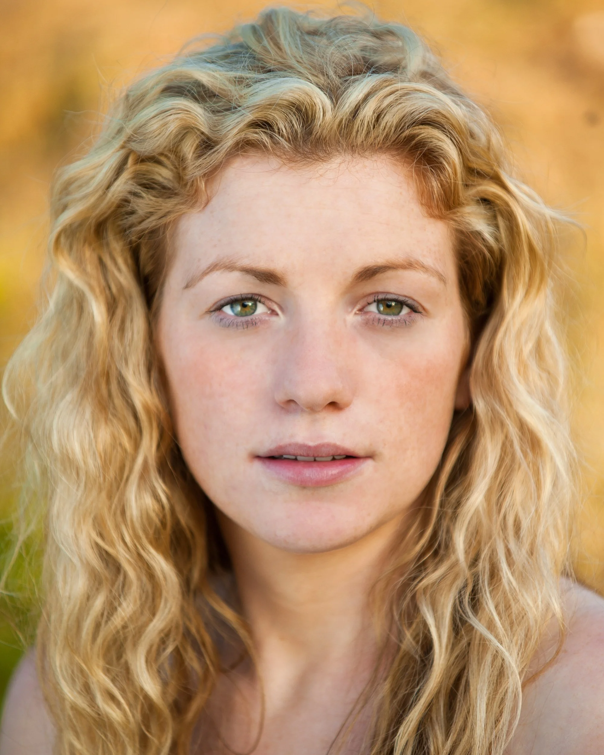 Outdoor portrait of a woman with curly blonde hair and green eyes surrounded by autumn foliage
