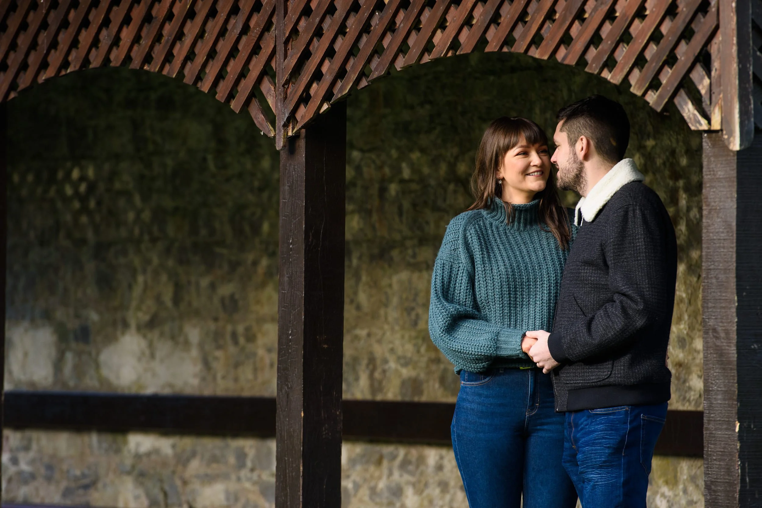 A couple standing under a wooden structure, smiling and holding hands, with a stone wall in the background at Coole Park Gort 